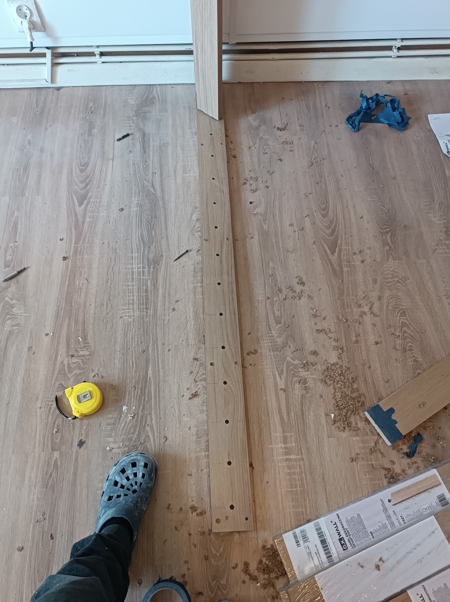 Przygotowanie do montażu paneli ściennych: nawiercona listwa z otworami na podłodze, obok miarka, wkręty i paczka nowych paneli.