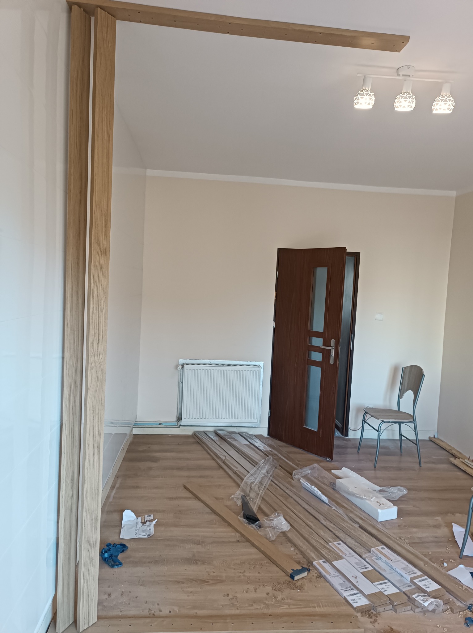 Montaż drewnianej ramy drzwiowej w jasnym pokoju, widoczne listwy przypodłogowe, grzejnik i drzwi z szybą, częściowo rozpakowane elementy ramy na podłodze.