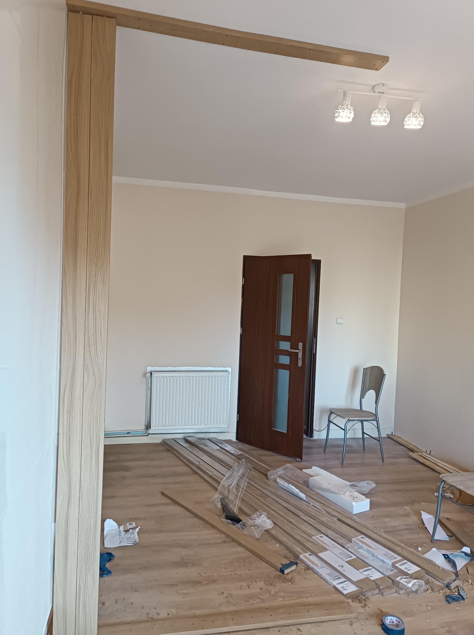 Montaż drewnianej konstrukcji na suficie i ścianie w trakcie remontu mieszkania, widoczne listwy przypodłogowe na podłodze, grzejnik i drzwi w tle.