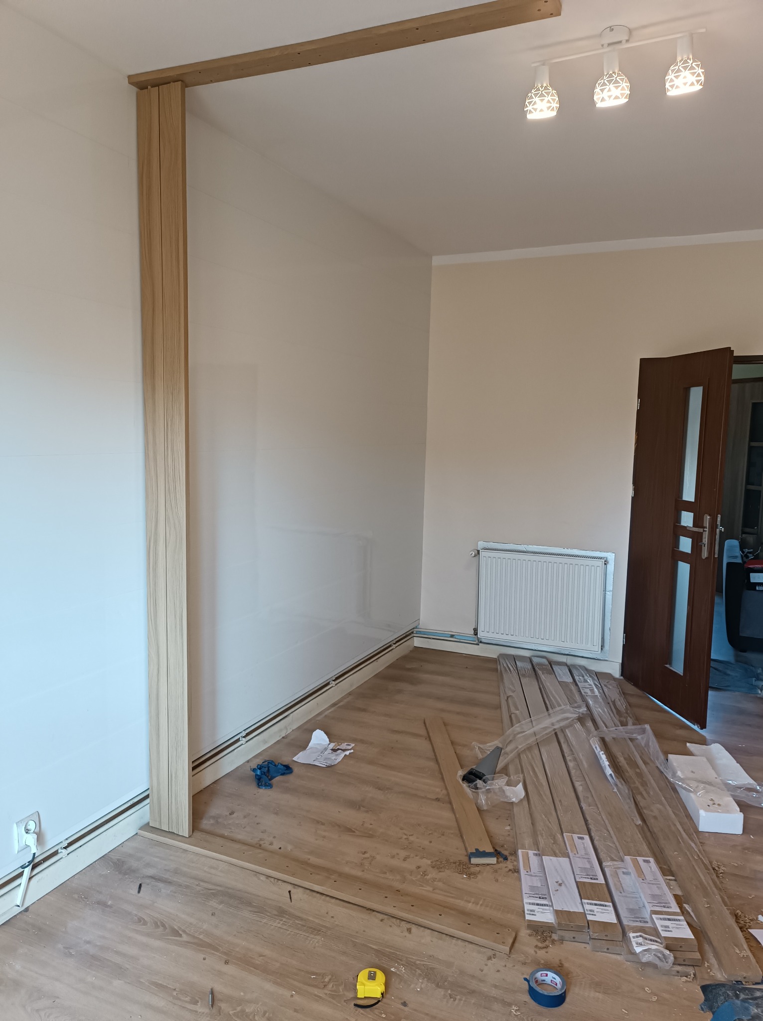 Montaż drewnianej konstrukcji w jasnym pokoju, widoczne elementy pionowe i poziome ramy, narzędzia, listwy przypodłogowe i częściowo zapakowane elementy wykończeniowe na podłodze.