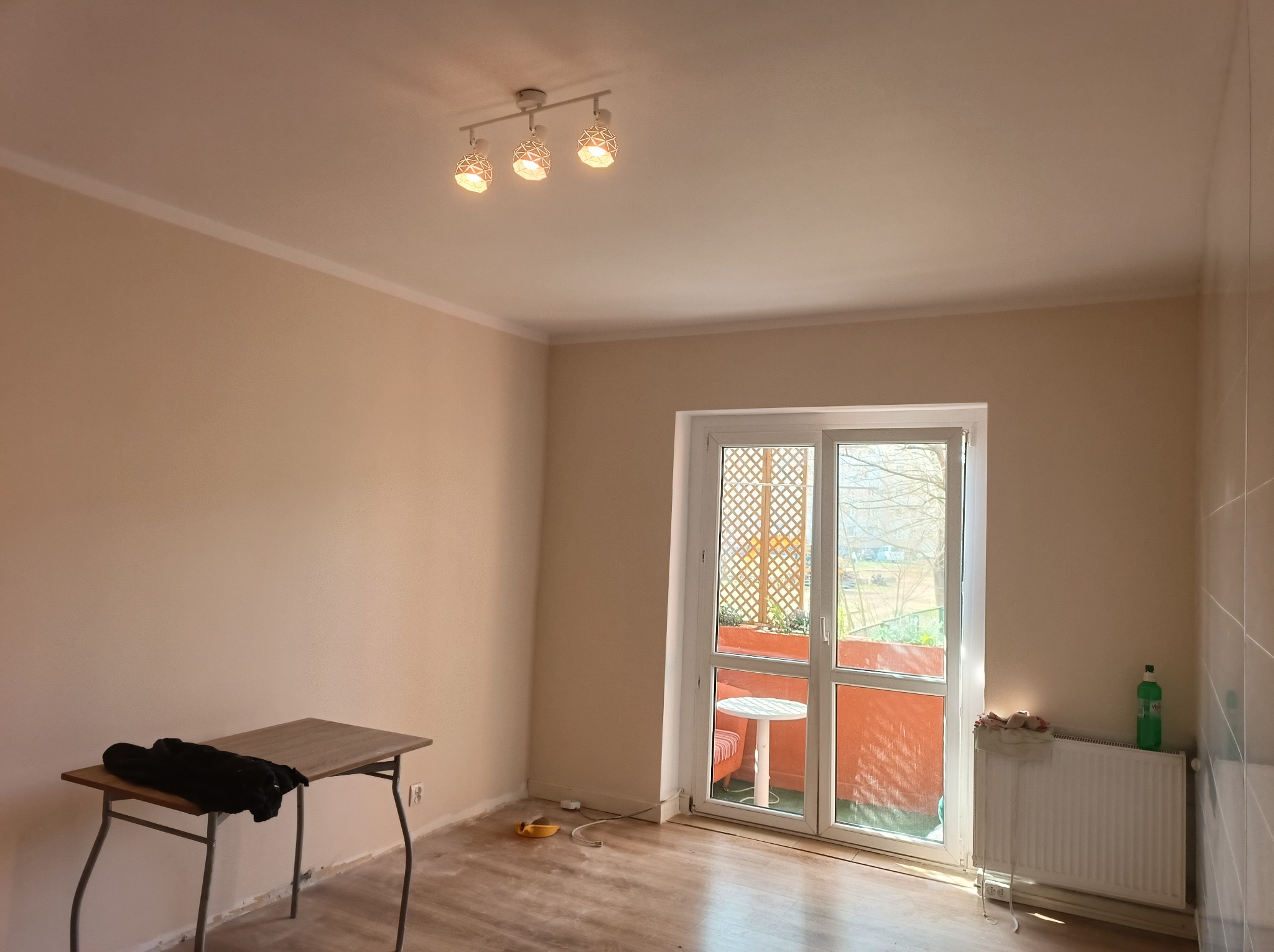 Pomalowane na beżowo puste pomieszczenie z balkonem, widoczny stolik, grzejnik z butelką płynu do czyszczenia i lampa sufitowa z trzema kloszami.