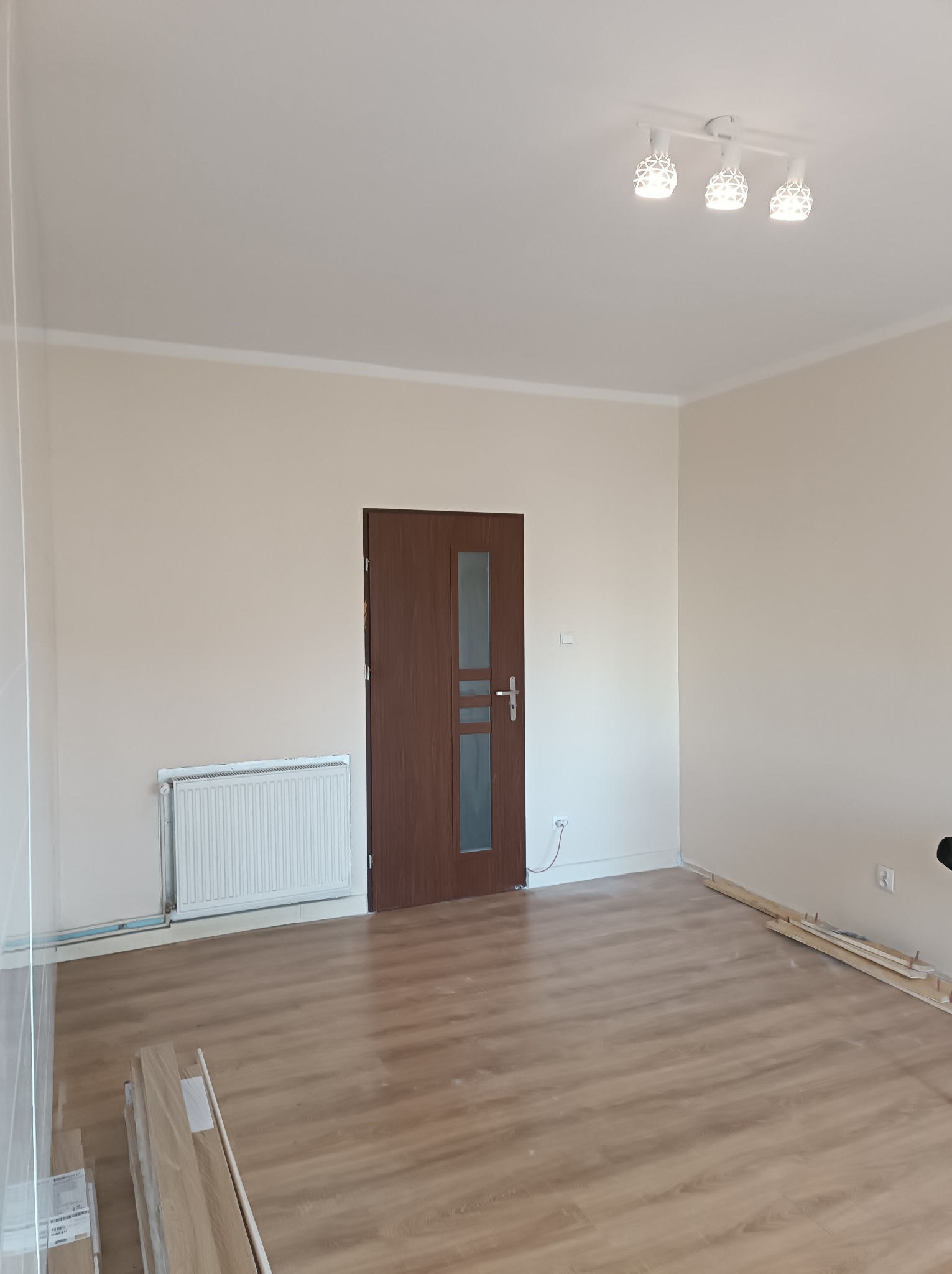 Puste pomieszczenie po remoncie z nową podłogą z paneli, świeżo pomalowanymi ścianami w odcieniach beżu, białym grzejnikiem i brązowymi drzwiami z szybą, oświetlone trzypunktową lampą sufitową.