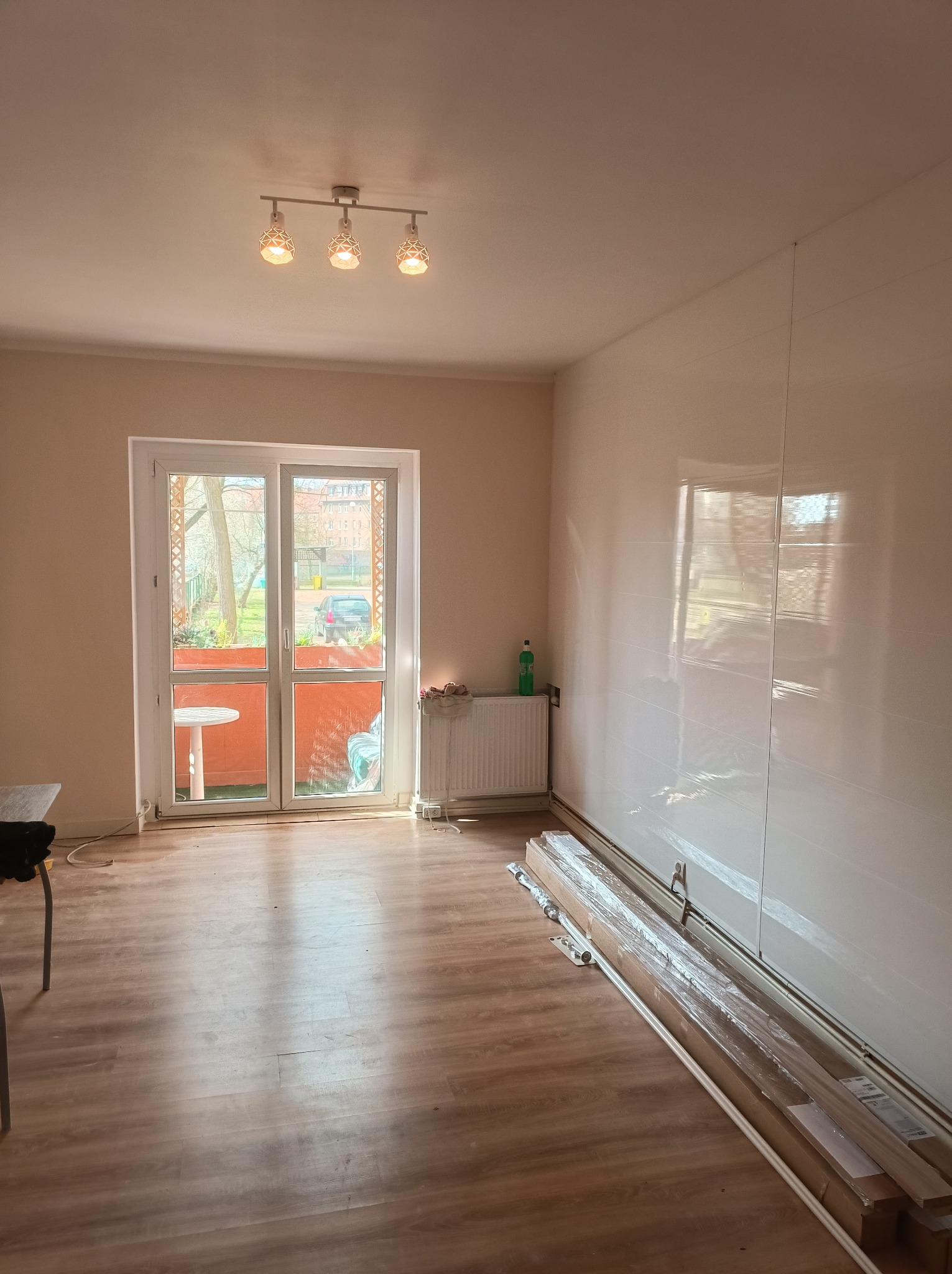 Pokój po remoncie z błyszczącymi białymi panelami ściennymi, drewnianą podłogą i oknem balkonowym wychodzącym na zewnątrz, z widocznymi listwami przypodłogowymi w opakowaniach.