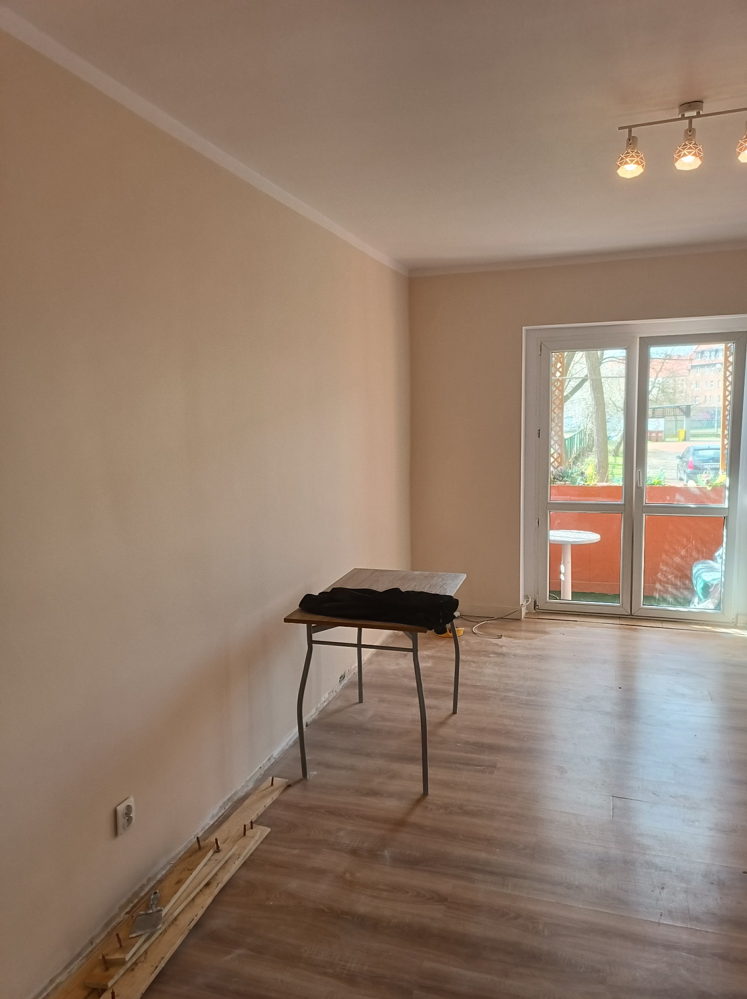 Pokój w trakcie prac wykończeniowych: świeżo pomalowane ściany w odcieniach beżu, listwa przypodłogowa częściowo zamontowana, widoczny stół warsztatowy i narzędzia.