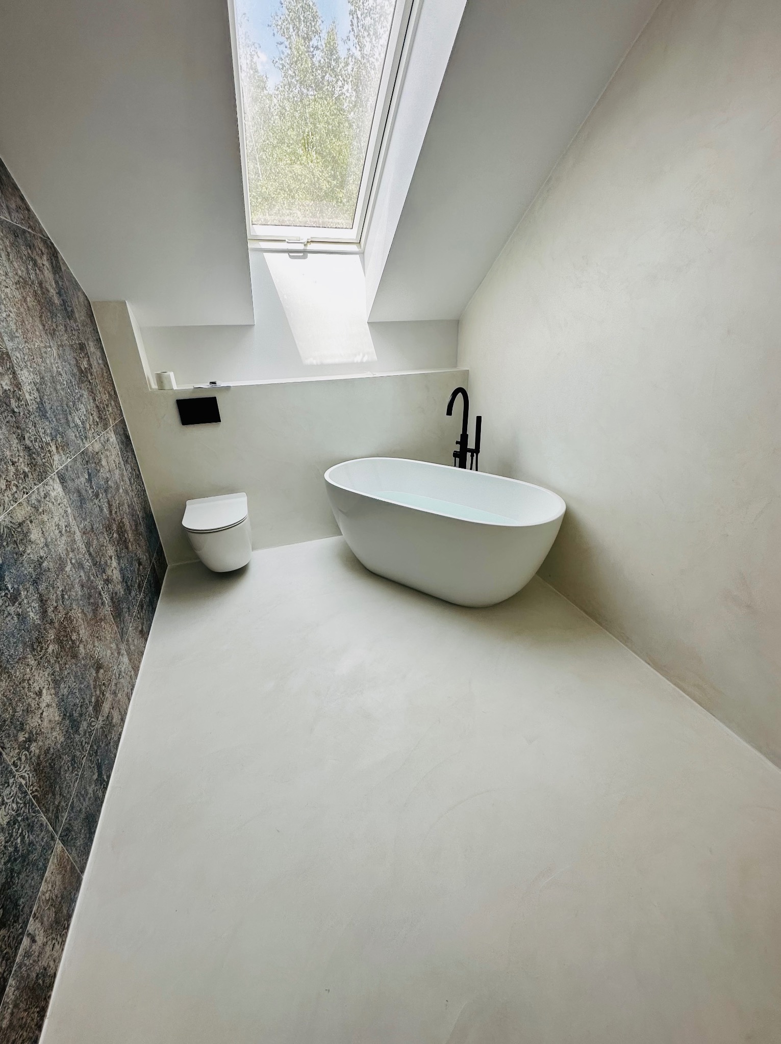 Minimalistyczna łazienka z białymi ścianami i podłogą, wolnostojąca wanna z czarną armaturą, toaleta podtynkowa i okno dachowe w skosie.