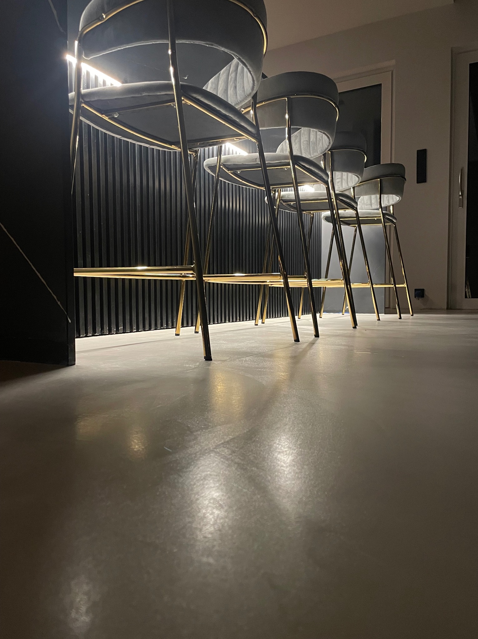 Szara posadzka z widocznymi refleksami światła, na której stoją cztery nowoczesne hokery z szarymi siedziskami i złotymi nogami, tło stanowi czarna ściana z pionowymi panelami.