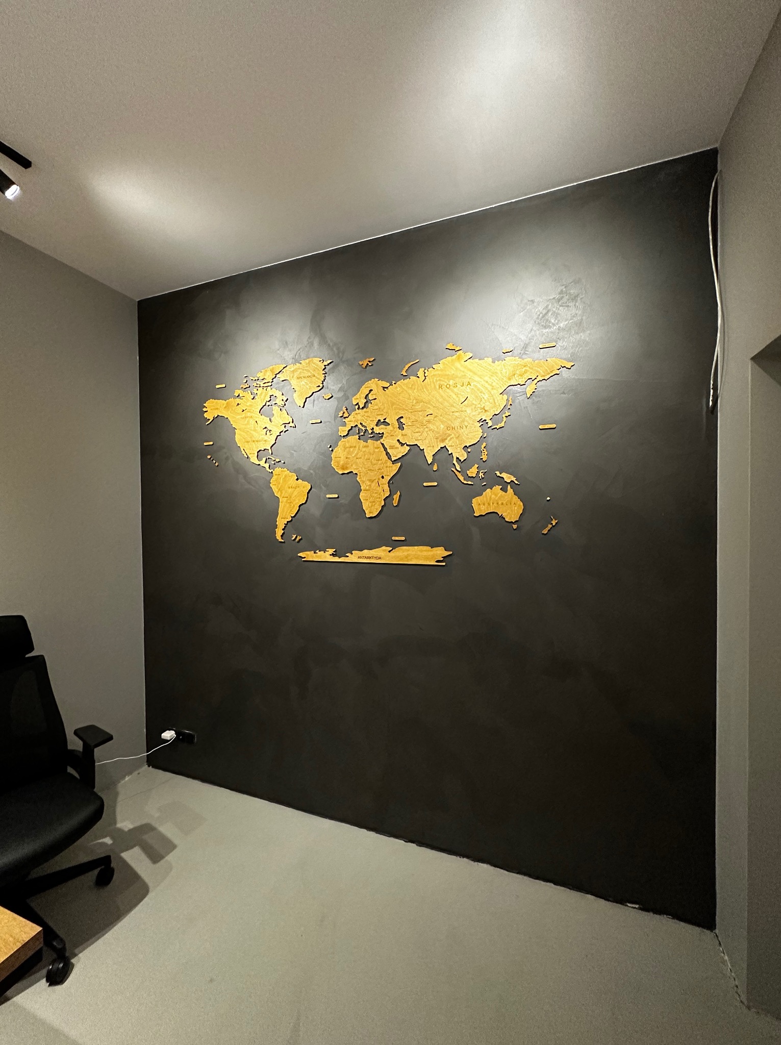 Ściana w biurze pokryta ciemnoszarym tynkiem dekoracyjnym z drewnianą mapą świata, oświetlona punktowo.