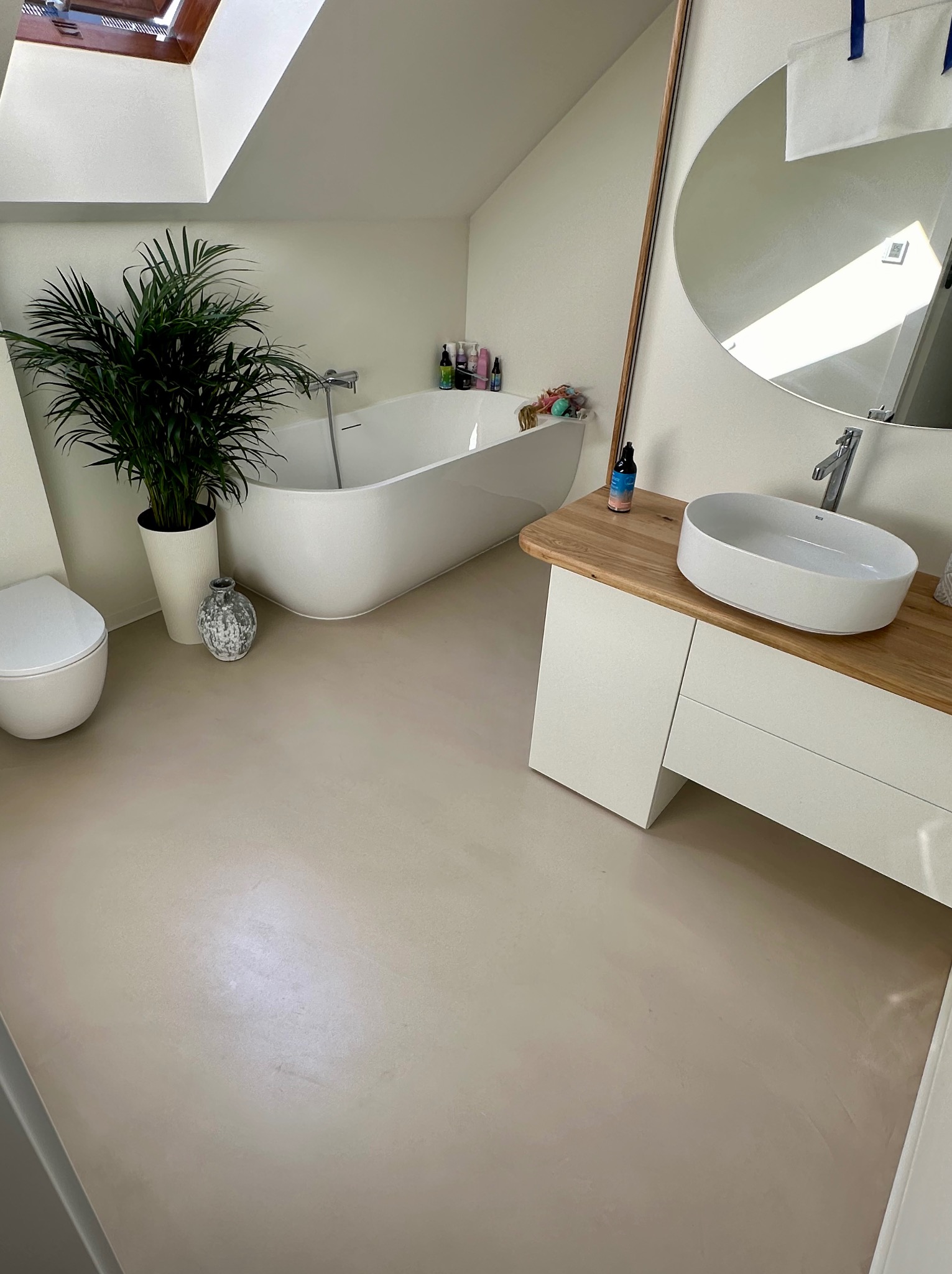 Jasna łazienka z podłogą w odcieniach beżu, wolnostojąca wanna, umywalka nablatowa na drewnianym blacie i duża roślina doniczkowa obok toalety.
