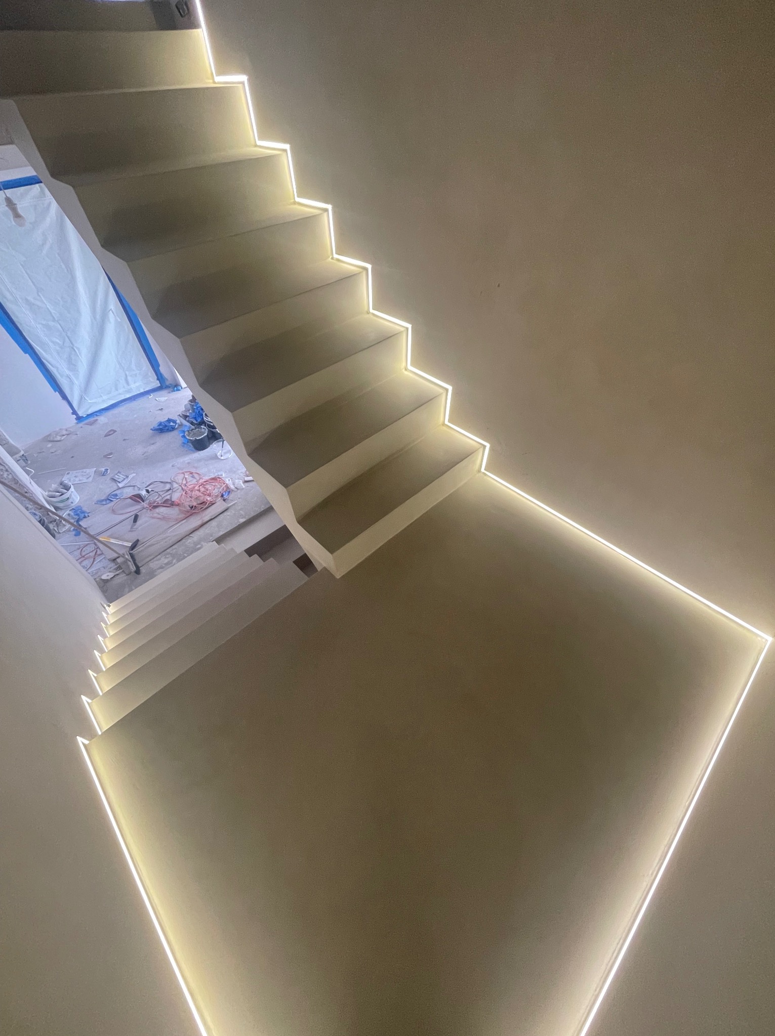 Schody wykończone jasnym materiałem z subtelnym oświetleniem LED wzdłuż krawędzi stopni, widok z góry na klatkę schodową w trakcie prac wykończeniowych.