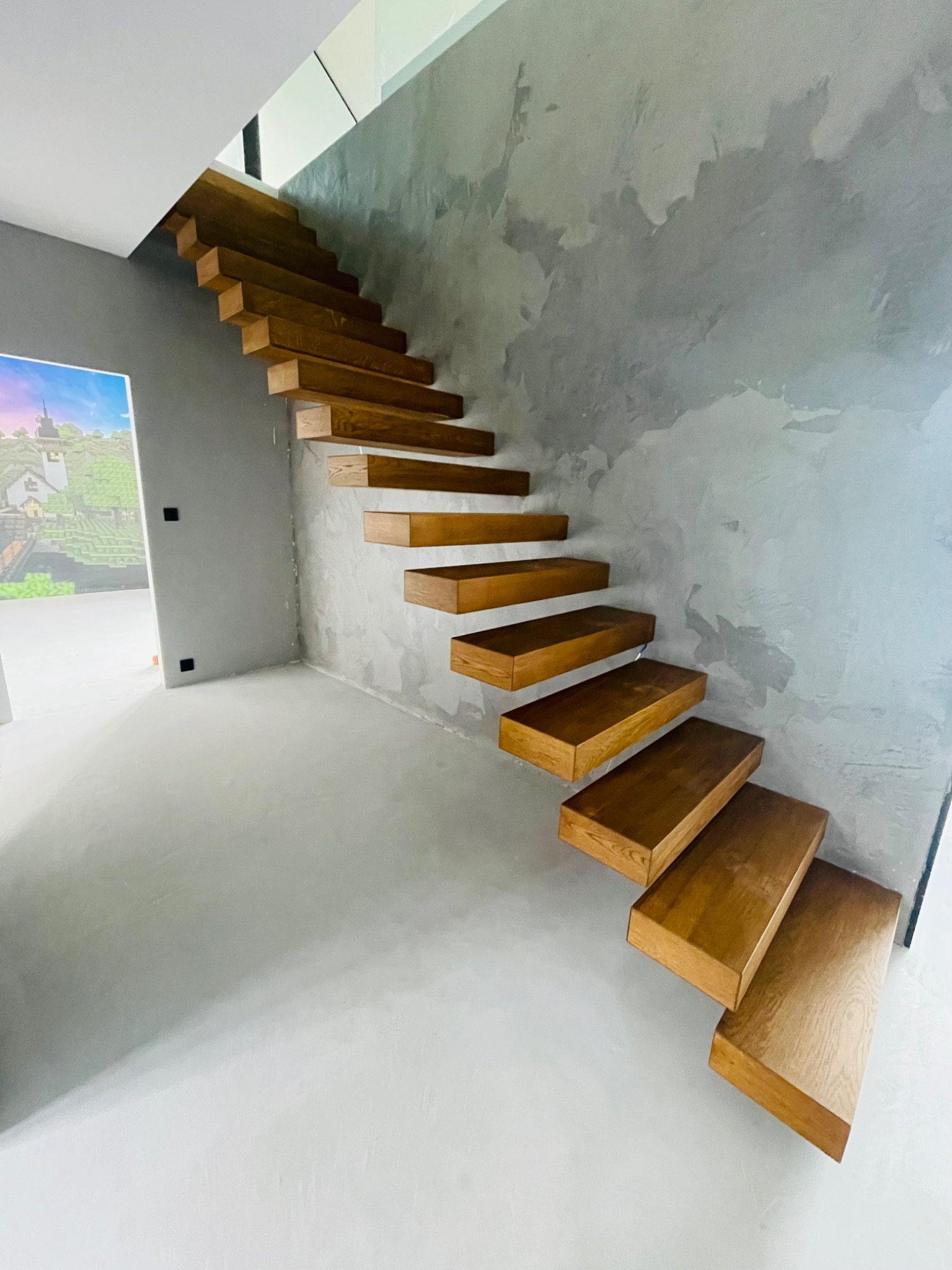 Nowoczesne, drewniane schody wspornikowe zamontowane przy ścianie z efektem betonu w jasnym wnętrzu z podłogą wykończoną w jasnym odcieniu szarości.