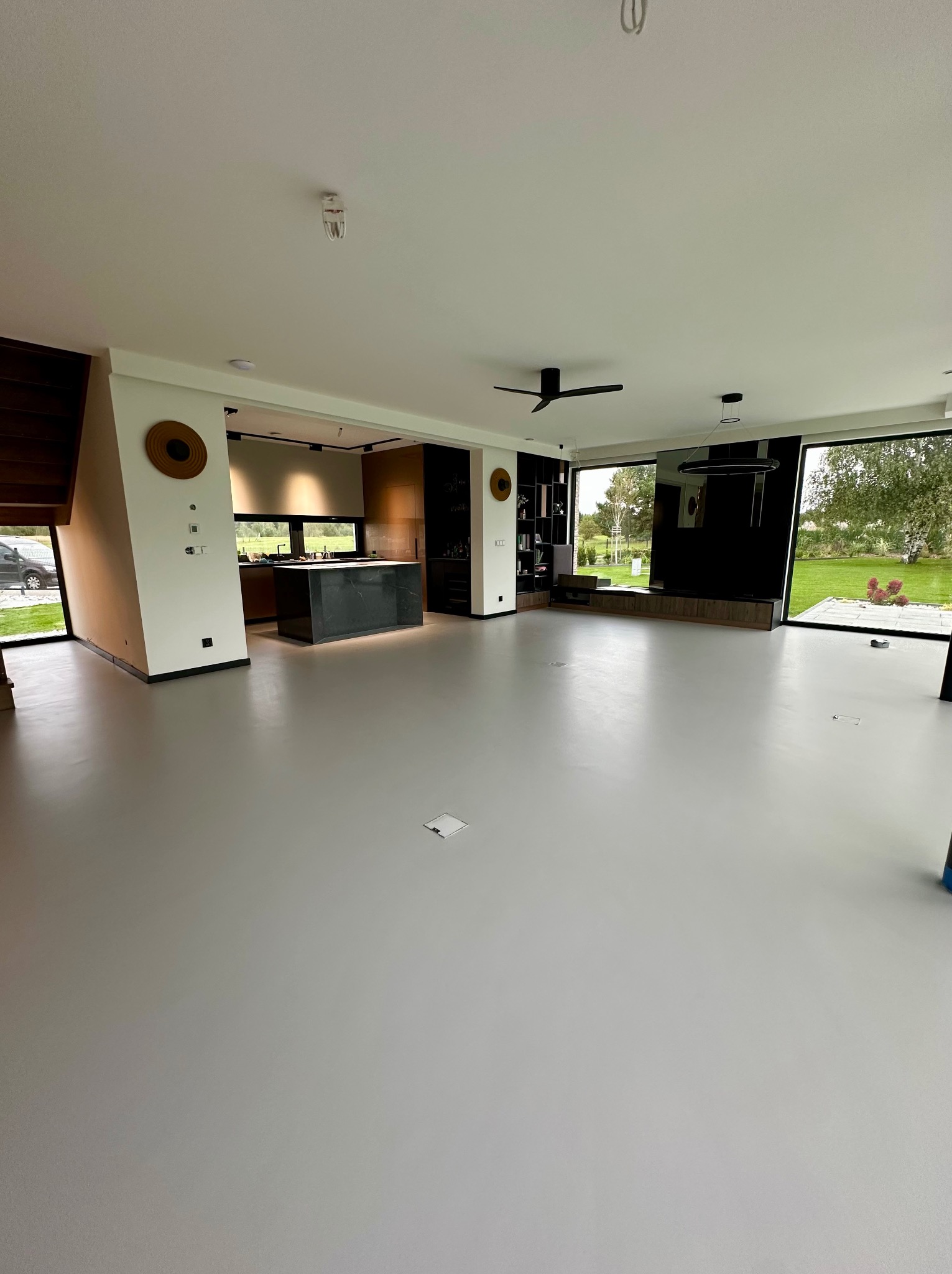 Jasnoszara posadzka z mikrocementu w przestronnym salonie z widokiem na ogród przez duże okna. Minimalistyczny design wnętrza z czarnymi akcentami i wyspą kuchenną.