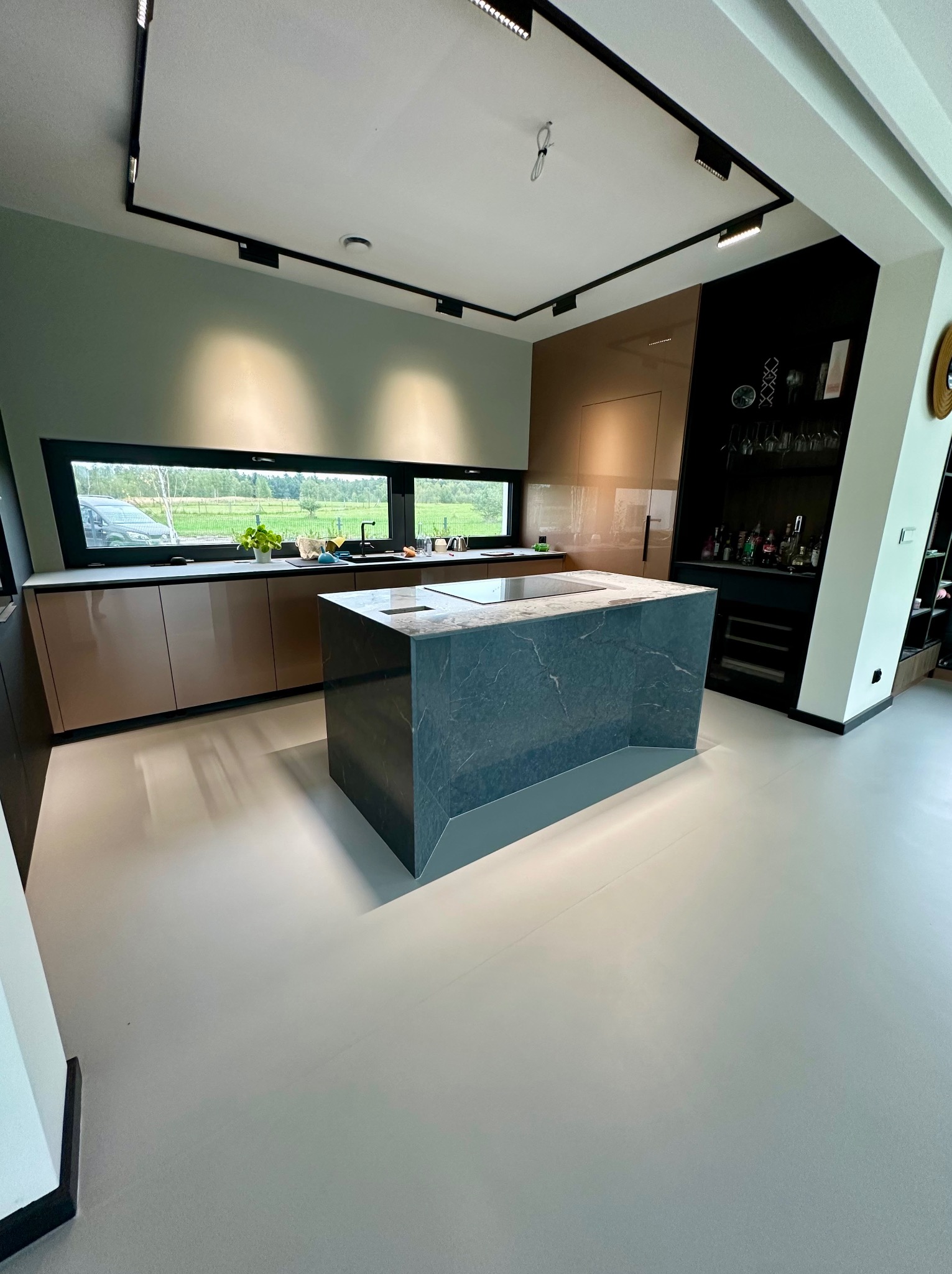 Nowoczesna kuchnia z wyspą o marmurowym wykończeniu i jasną posadzką. Minimalistyczne oświetlenie punktowe i panoramiczne okno z widokiem na zieleń.