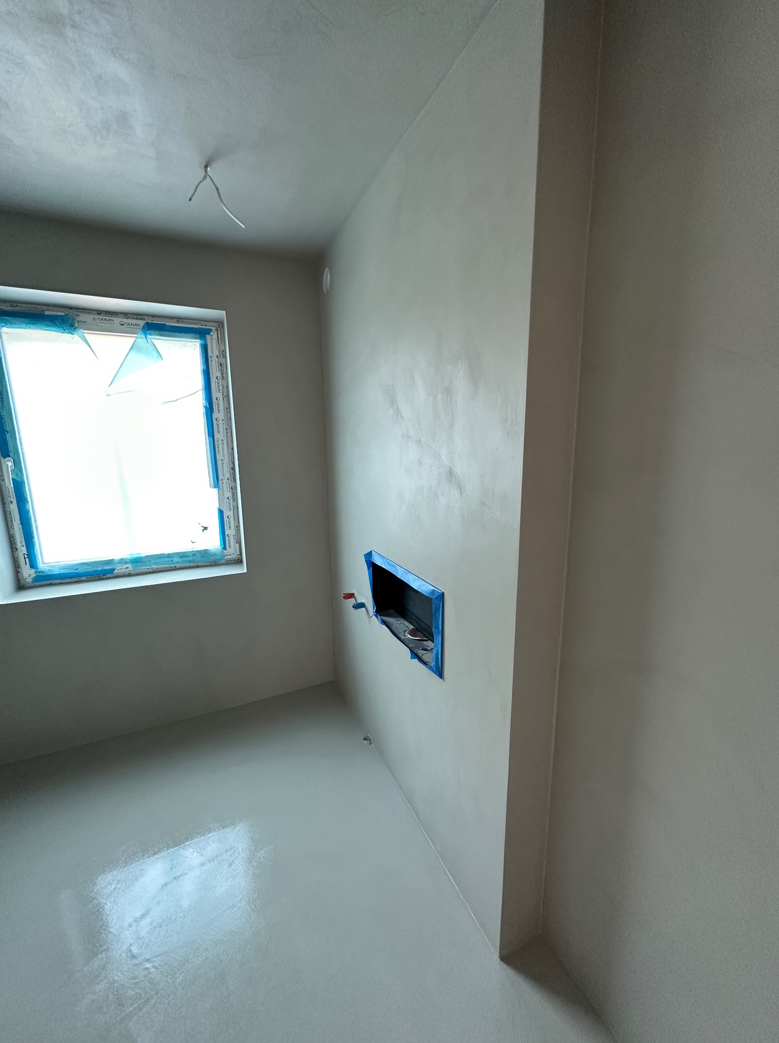 Jasne wnętrze po aplikacji dekoracyjnej powłoki na ścianach i podłodze. Okno zabezpieczone folią. Widoczny fragment instalacji w ścianie.