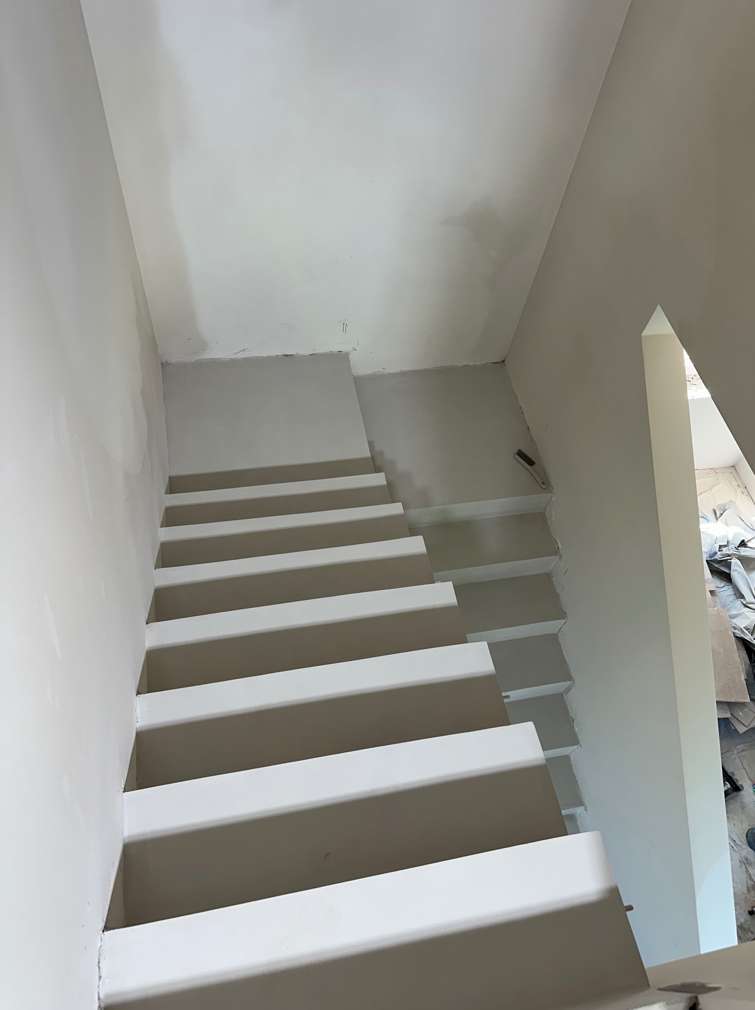 Wykończone schody wewnętrzne w jasnej tonacji, widok z góry. Ściany i sufit w trakcie prac wykończeniowych. Widoczny fragment narzędzia malarskiego.