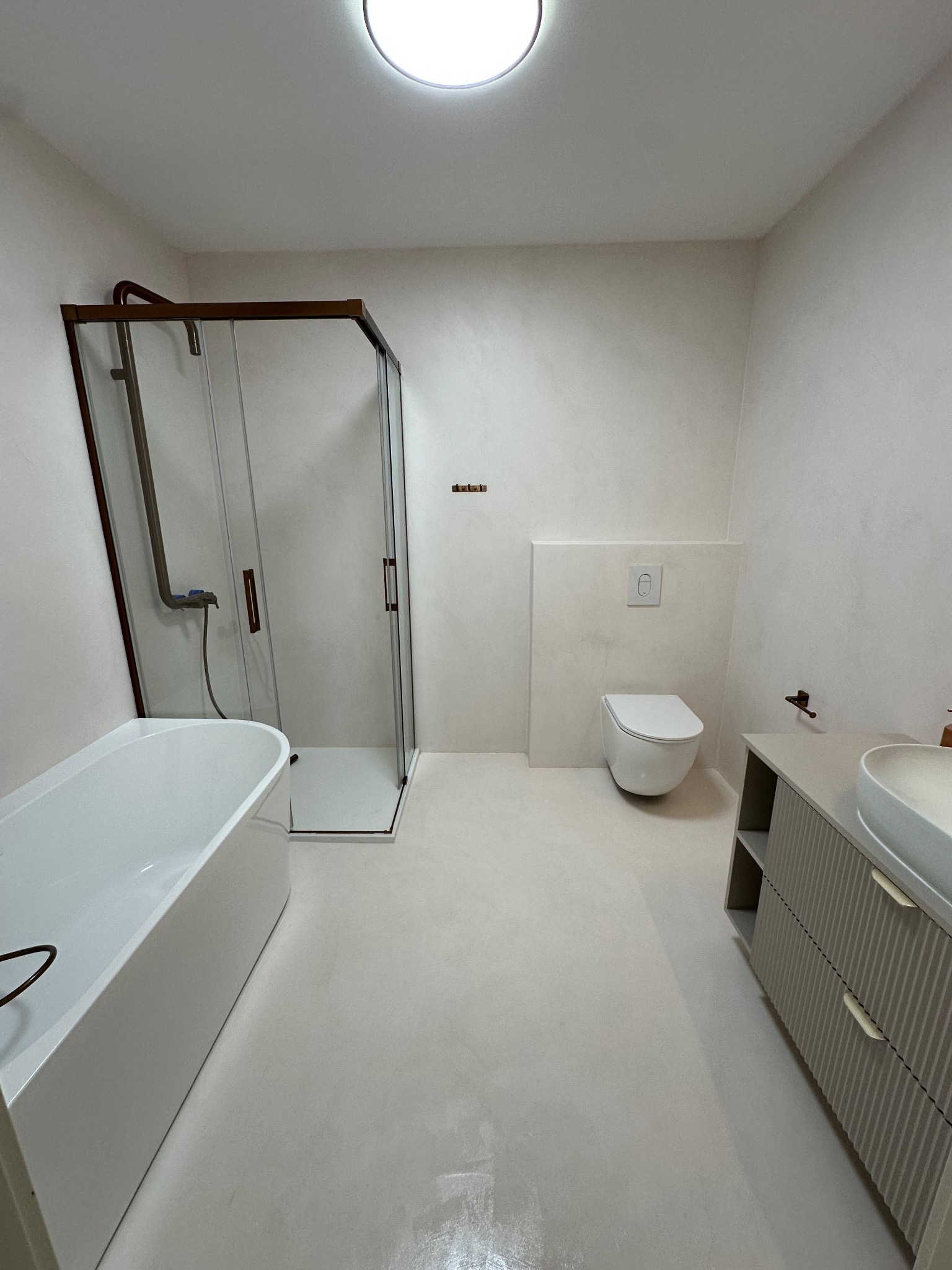 Jasna łazienka z kabiną prysznicową, wanną, umywalką z szafką i toaletą. Ściany i podłoga wykończone jednolitą, jasną powierzchnią. Minimalistyczny design.