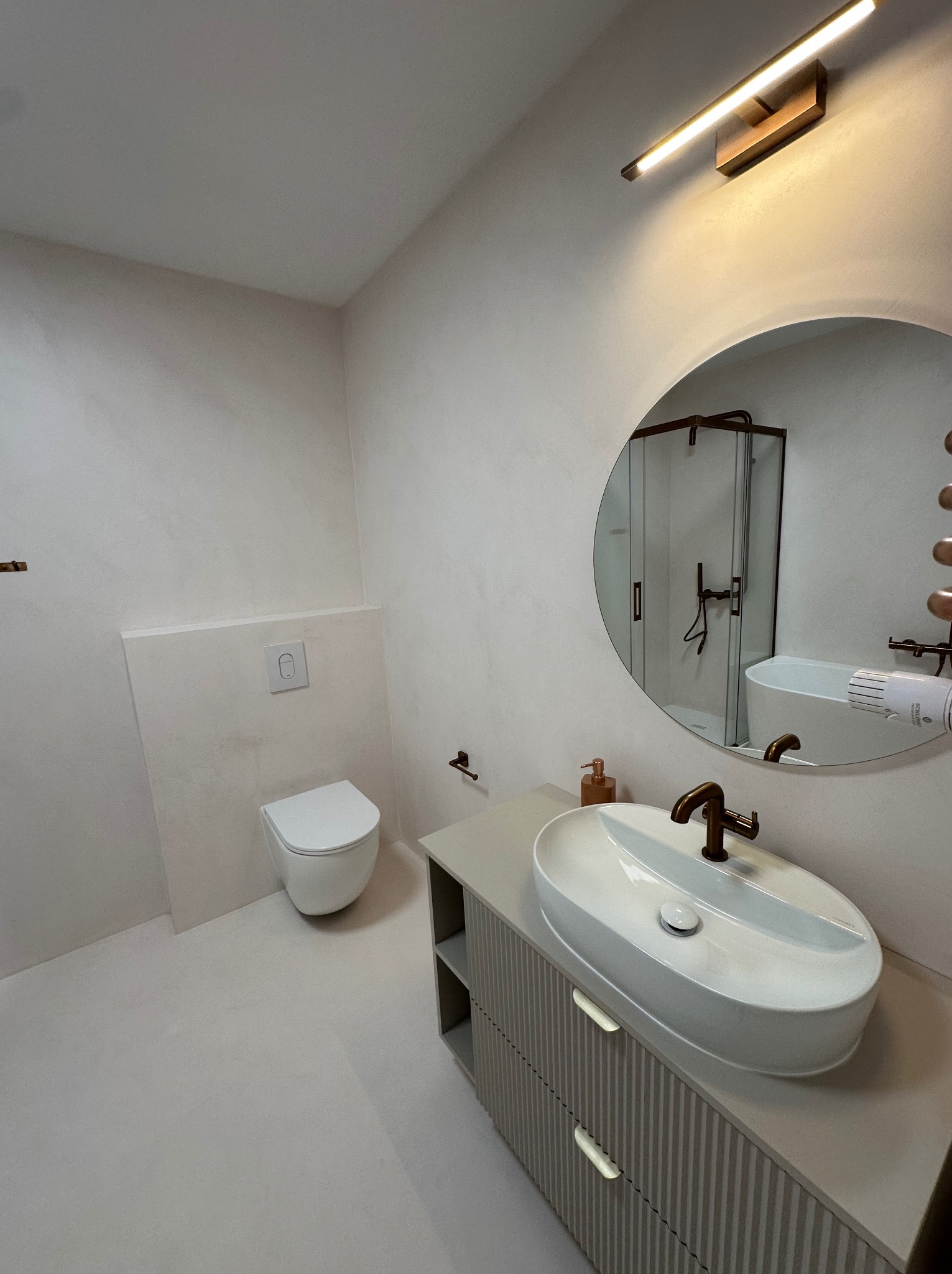 Jasna łazienka z toaletą, umywalką na szafce i okrągłym lustrem, wykończona jednolitą powierzchnią ścian i podłogi. Widoczna kabina prysznicowa w odbiciu lustra.