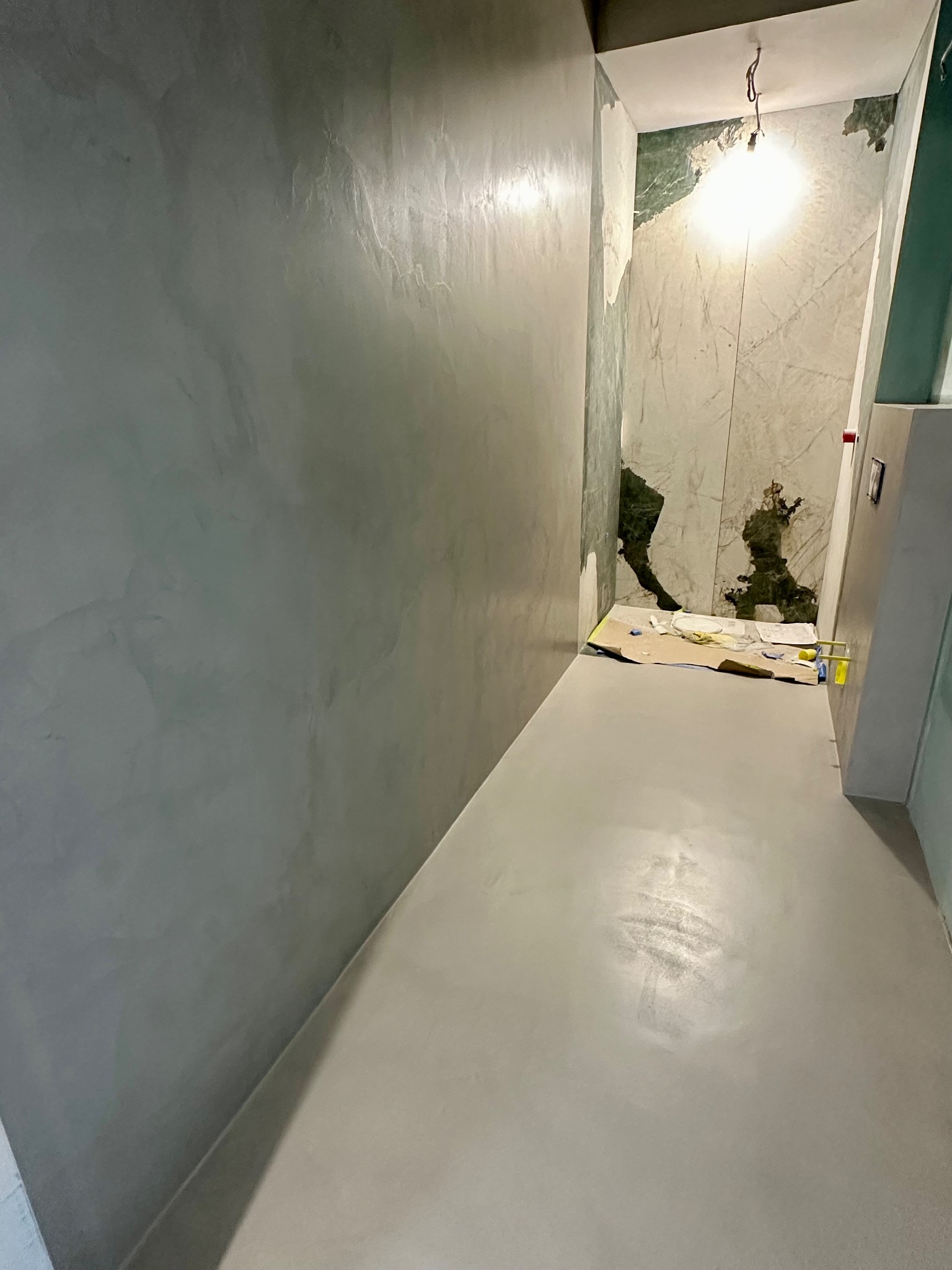 Szara, gładka posadzka i ściana w pomieszczeniu w trakcie prac wykończeniowych. Na podłodze karton zabezpieczający, widoczne narzędzia i niedokończona ściana.