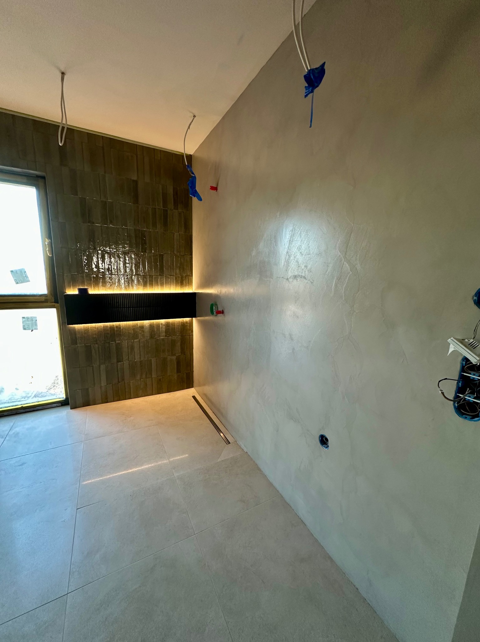 Wykończona łazienka z szarą, błyszczącą ścianą, płytkami imitującymi cegłę i podświetlaną szafką. Widoczne niedokończone instalacje elektryczne.