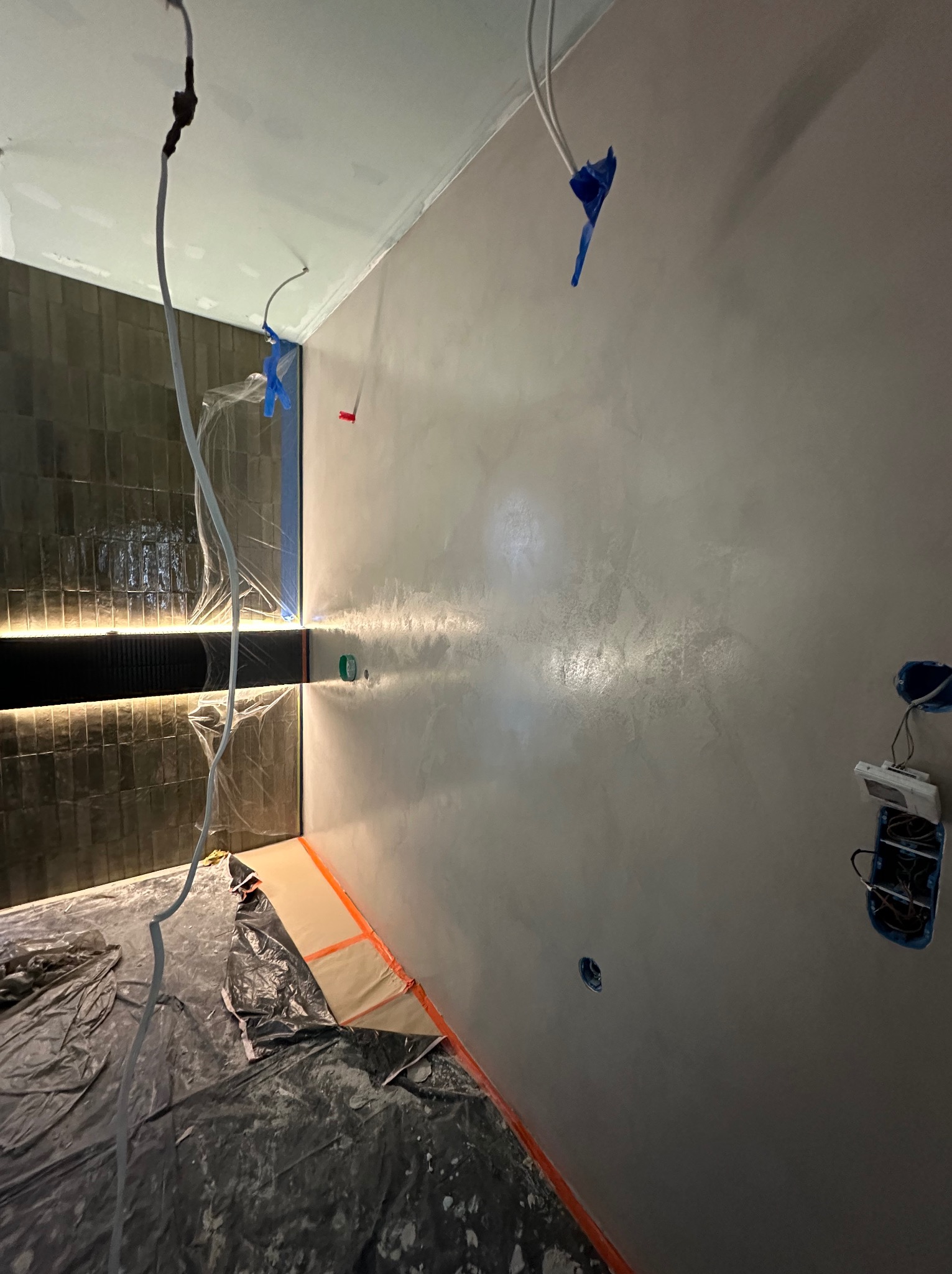 Ściana w trakcie wykończenia z widocznymi zabezpieczeniami i instalacją elektryczną. Szara, gładka powierzchnia z delikatnym połyskiem. Widoczne płytki na ścianie w tle.