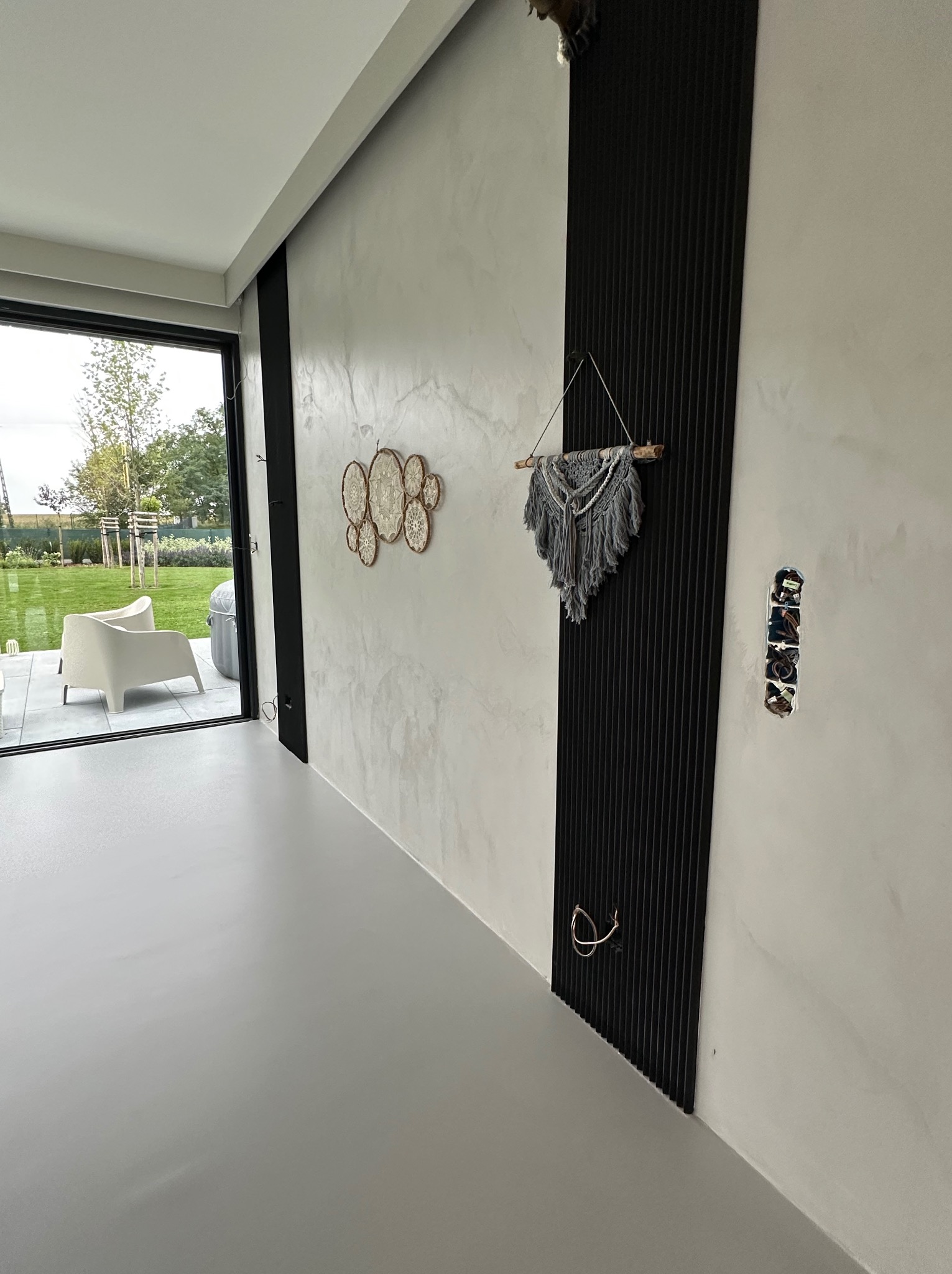 Jasnoszara posadzka w nowoczesnym wnętrzu z wyjściem na taras. Ściana z dekoracyjną strukturą, czarny panel z ozdobą i widoczne puszki elektryczne.