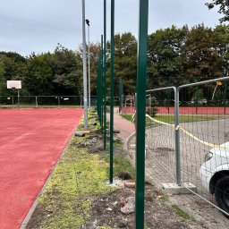 Vadym Hrebeniuk - Zielone, metalowe słupki ogrodzeniowe wkopane w ziemię wzdłuż czerwonego boiska sportowego. Widoczne tymczasowe ogrodzenie i fragment samochodu.