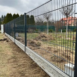 Vadym Hrebeniuk - Nowoczesne ogrodzenie panelowe z podmurówką betonową o fakturze kamienia, widok z perspektywy ziemi, na tle zieleni i domu z czerwoną dachówką.