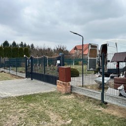 Vadym Hrebeniuk - Nowoczesne ogrodzenie panelowe z bramą wjazdową, szara kostka brukowa, skrzynka na murku z cegły, biały bus w tle, pochmurne niebo.