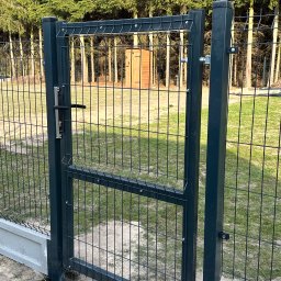 Vadym Hrebeniuk - Nowoczesna furtka z paneli metalowych w kolorze grafitowym, z widoczną klamką i zawiasami, na tle zieleni drzew i trawy. Solidna konstrukcja z podmurówką.