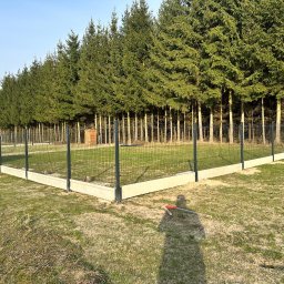 Vadym Hrebeniuk - Nowoczesne ogrodzenie panelowe z podmurówką betonową, otoczone zielenią i drzewami iglastymi. Widoczny cień osoby i narzędzia, sugerujące trwający montaż.