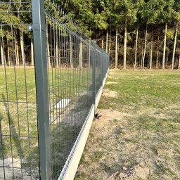 Vadym Hrebeniuk - Nowoczesne ogrodzenie panelowe z podmurówką w Lublinie, widok z perspektywy gruntu, z lasem w tle. Szara kolorystyka paneli i słupków.