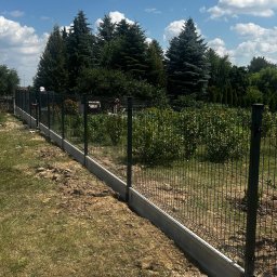 Vadym - Ciemnozielone ogrodzenie panelowe z betonową podmurówką, częściowo zamontowane na nierównym terenie, z bujną zielenią i sosnami w tle pod błękitnym niebem.