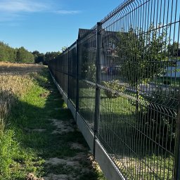 Vadym - Długie, ciemnoszare ogrodzenie panelowe z betonową podmurówką, rozciągające się wzdłuż pola zboża i posesji mieszkalnej, pod bezchmurnym, błękitnym niebem.