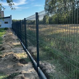 Vadym - Nowo zamontowane ciemnoszare ogrodzenie panelowe z betonową podmurówką, ciągnące się wzdłuż posesji, oddzielające trawnik od pola z wysoką trawą. W tle nowoczesny dom i drzewa pod błękitnym niebem.