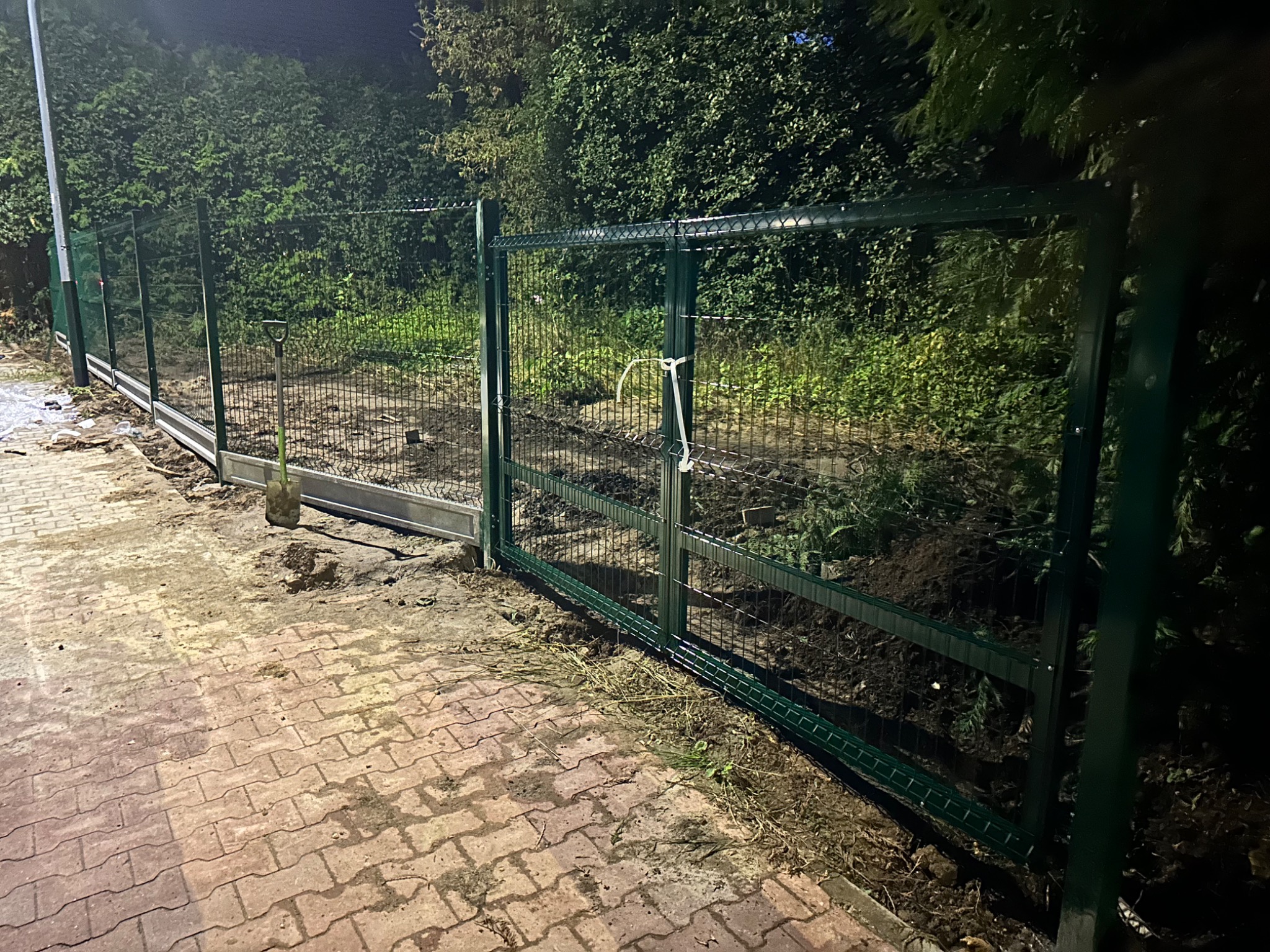 Montaż zielonego ogrodzenia panelowego z bramą przesuwną, widoczna łopata i rozkopana ziemia, kostka brukowa. Ujęcie wieczorne.
