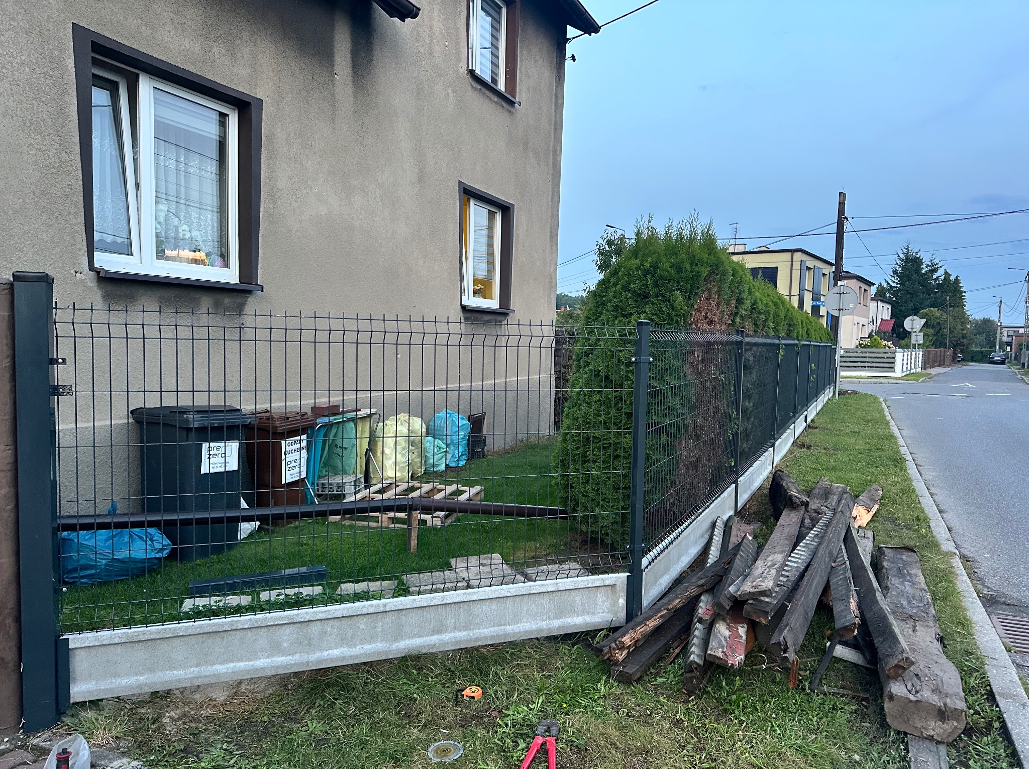 Ciemny panel ogrodzeniowy z podmurówką, widok z boku domu. Na trawie leżą narzędzia i drewniane elementy. W tle zieleń i budynki.