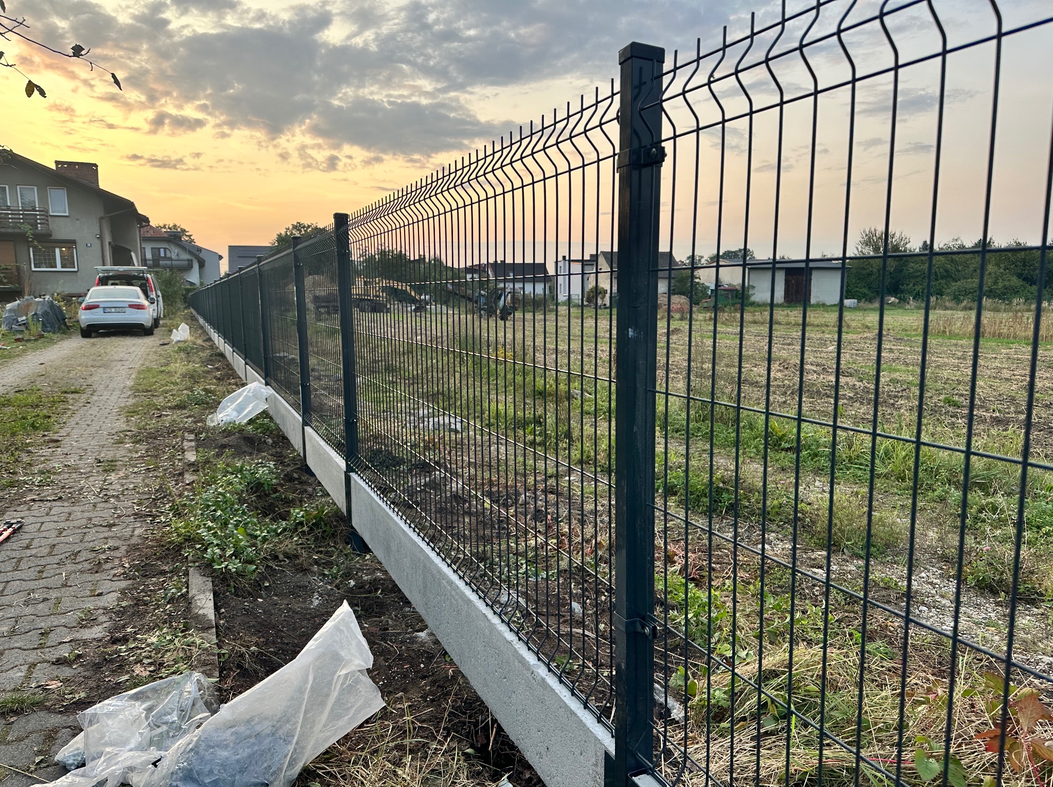 Nowoczesne ogrodzenie panelowe z podmurówką betonową, widok z boku, w tle dom jednorodzinny i zachodzące słońce.
