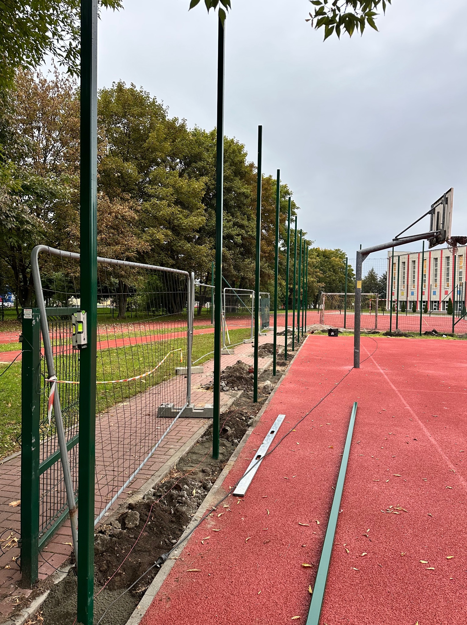 Montaż ogrodzenia metalowego na boisku: widoczne słupki, wykop, poziomica i fragment boiska z koszem. Etap prac przed finalnym zamontowaniem paneli.