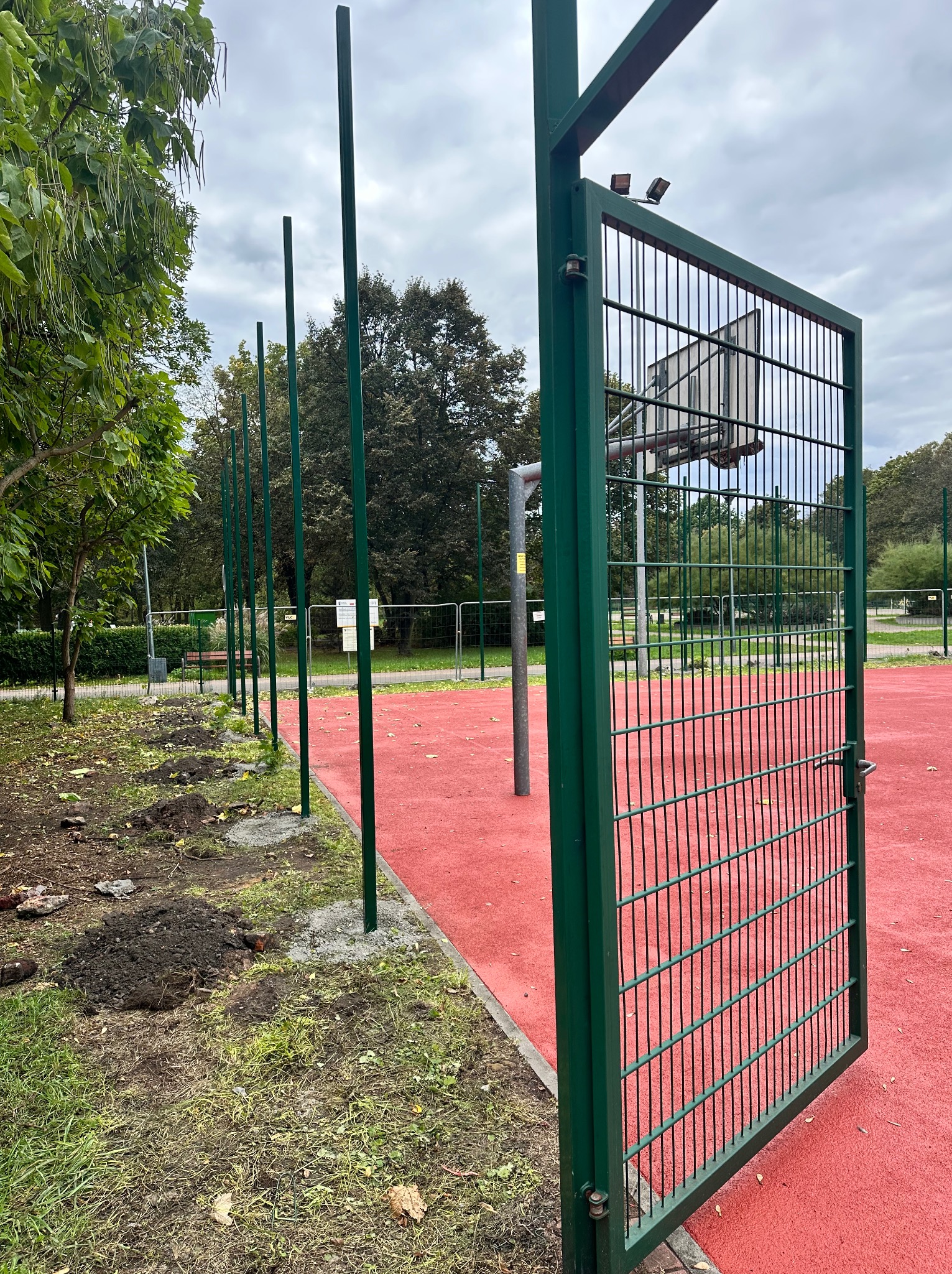 Otwarta, zielona metalowa brama na czerwonym boisku. Widoczne słupki ogrodzenia w trakcie montażu, z wykopanymi dołami. W tle zieleń parku i pochmurne niebo.