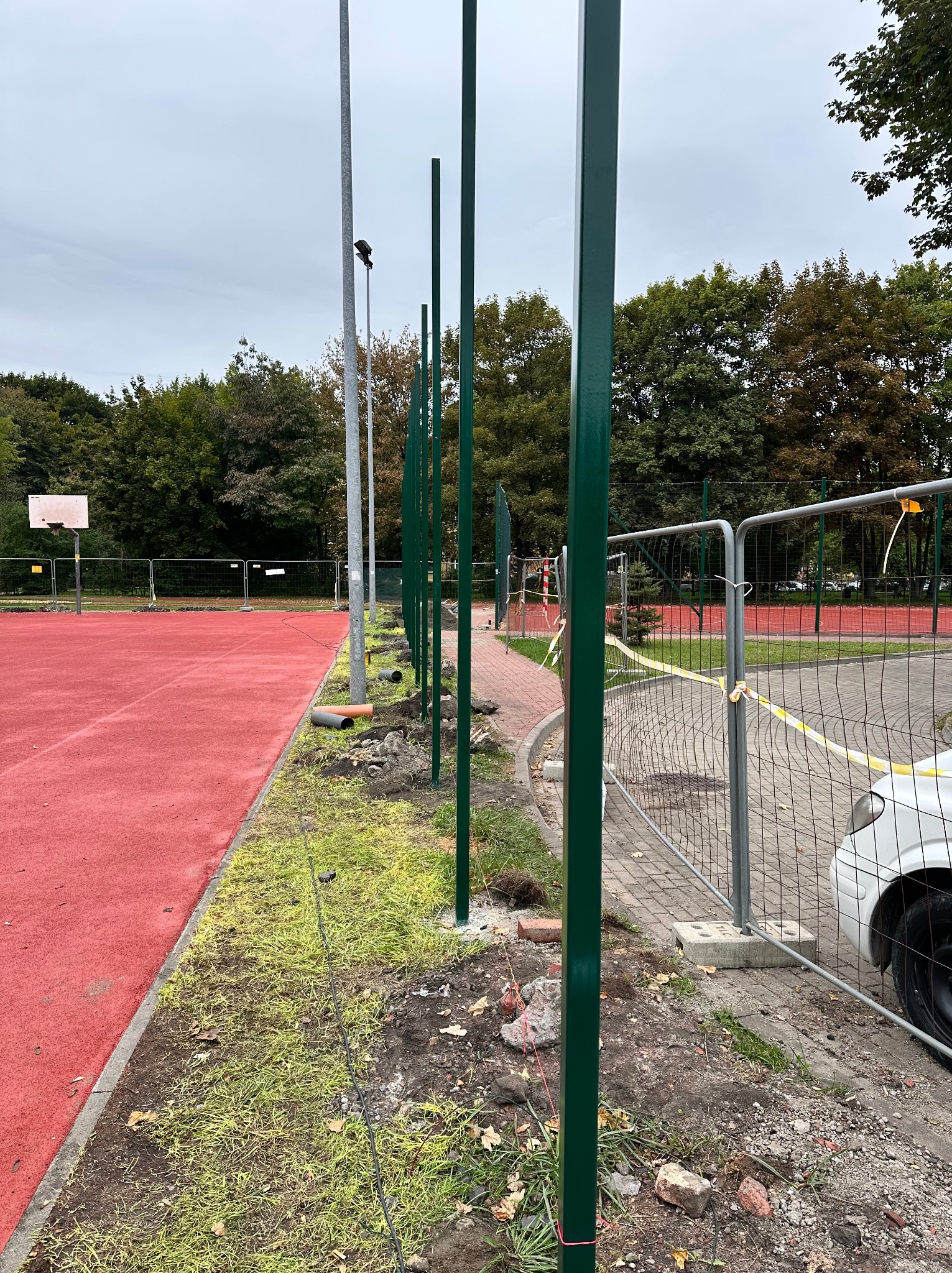 Zielone, metalowe słupki ogrodzeniowe wkopane w ziemię wzdłuż czerwonego boiska sportowego. Widoczne tymczasowe ogrodzenie i fragment samochodu.