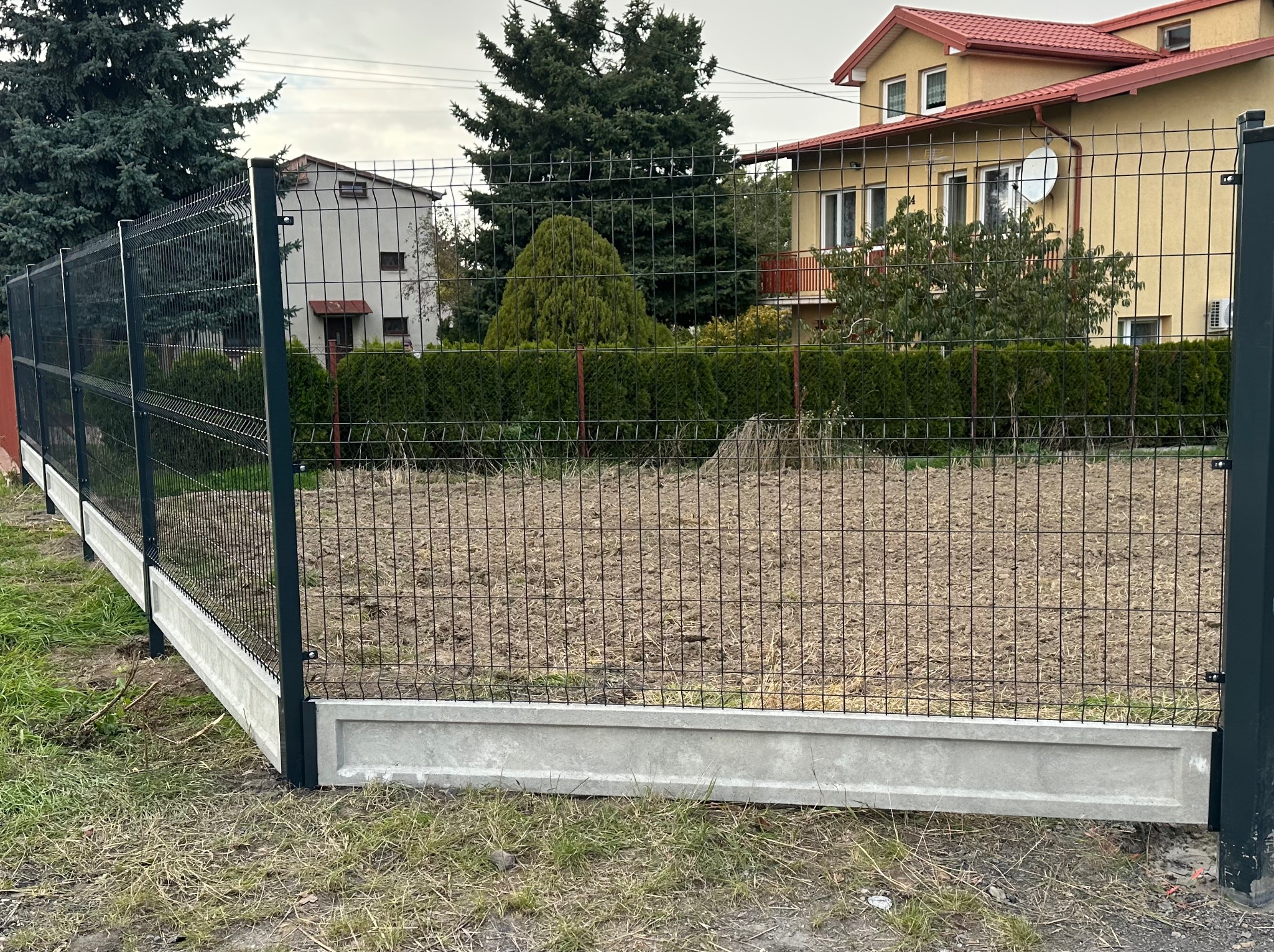 Nowoczesne ogrodzenie panelowe z podmurówką betonową, widok z niskiej perspektywy. Ciemne panele na tle zieleni drzew i zabudowań.