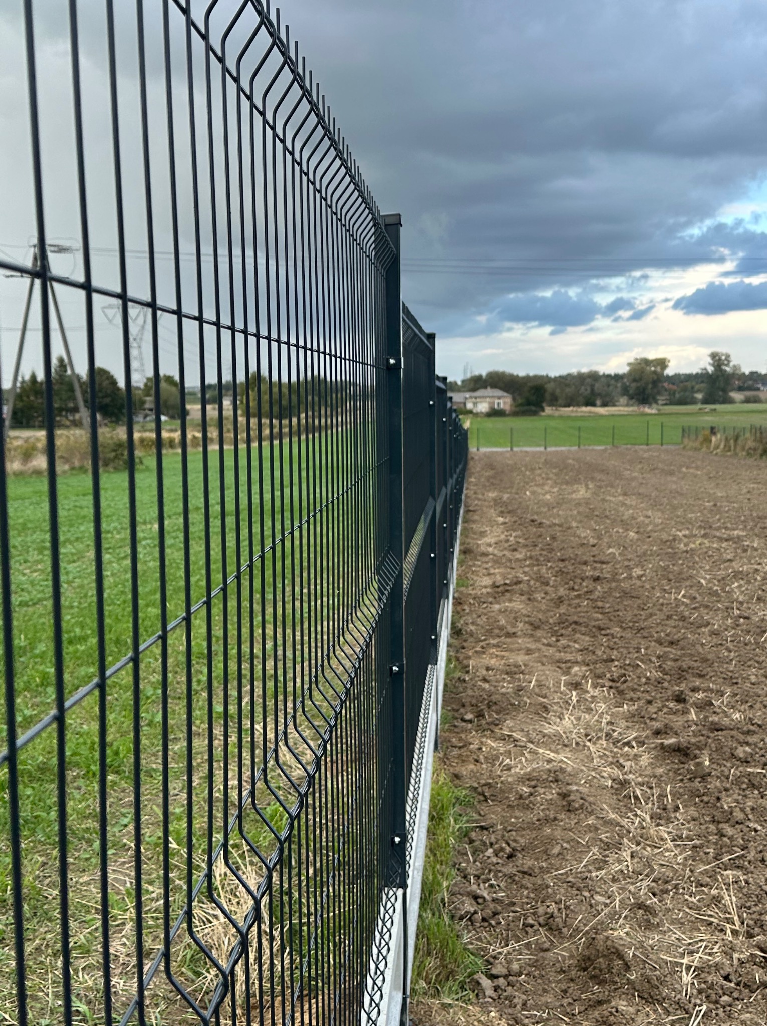 Nowoczesne ogrodzenie panelowe w kolorze grafitowym, oddzielające zielony teren od świeżo zaoranej ziemi, w tle pochmurne niebo i budynek.
