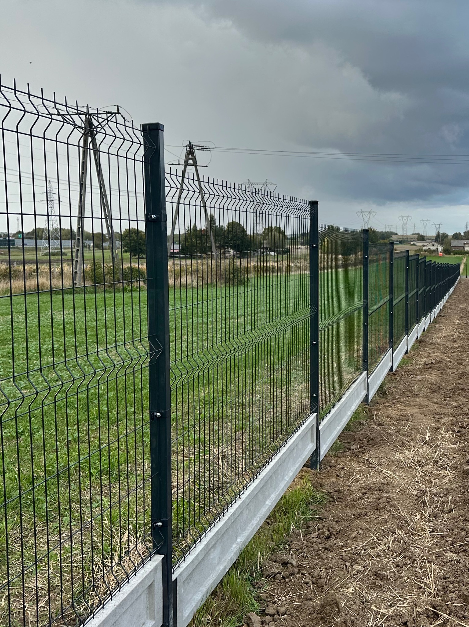 Długi, panelowy płot z podmurówką, ciągnący się wzdłuż zielonego pola pod pochmurnym niebem. Widoczne słupy energetyczne w oddali.