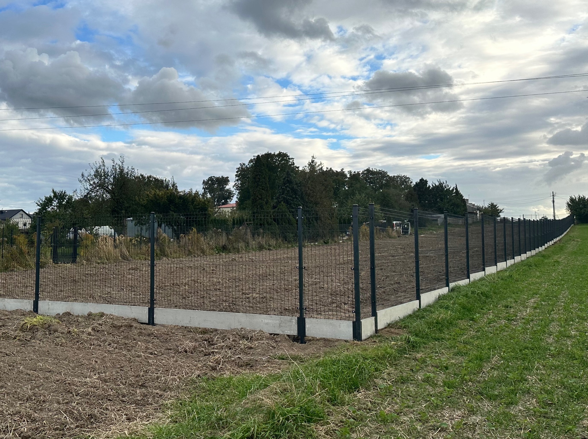 Długi, nowy panelowy płot z podmurówką, rozciągający się wzdłuż pola, z widocznymi słupkami i siatką. W tle drzewa i pochmurne niebo.