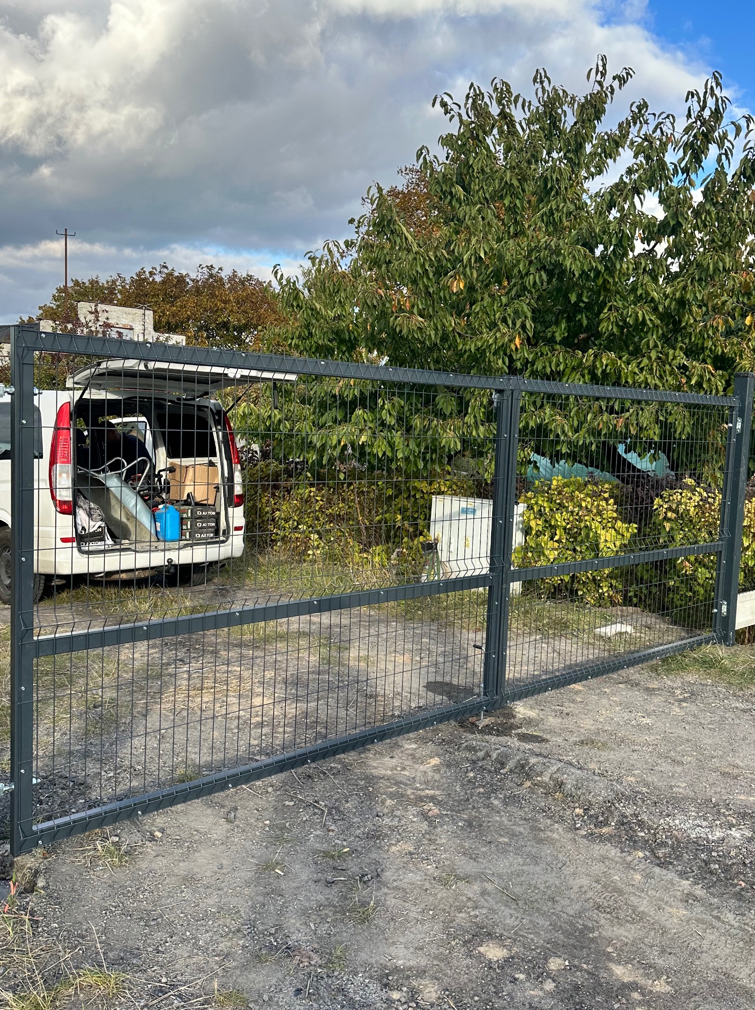 Nowoczesna, szara brama panelowa na posesji, z otwartym bagażnikiem białego samochodu dostawczego w tle, sugerująca świeży montaż.