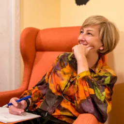 Uśmiechnięta kobieta z krótkimi blond włosami siedzi w pomarańczowym fotelu, trzymając kartkę i niebieski długopis, spoglądając w górę z zamyśleniem, w tle abstrakcyjna dekoracja ścienna.