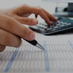 Obliczanie danych finansowych: ręka z długopisem nad arkuszem kalkulacyjnym, w tle kalkulator i monitor. Precyzyjne wyliczenia i analiza danych.