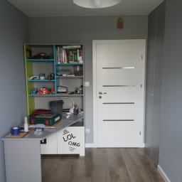 SinggaDruk Filip Sinulingga - Sztuka na każdej ścianie - Szary pokój dziecięcy po malowaniu: biurko z regałem, białe drzwi z poziomymi paskami, drewniana podłoga. Na ścianie dekoracja w kształcie głowy Buddy.