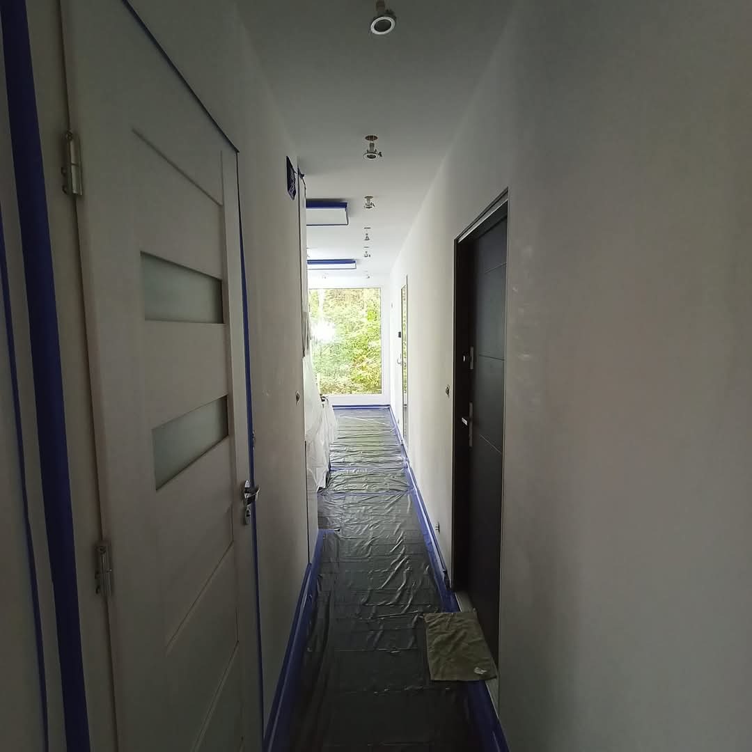 Korytarz w trakcie malowania: ściany świeżo pomalowane na biało, podłoga zabezpieczona folią, drzwi oklejone taśmą malarską. Widok w głąb korytarza.