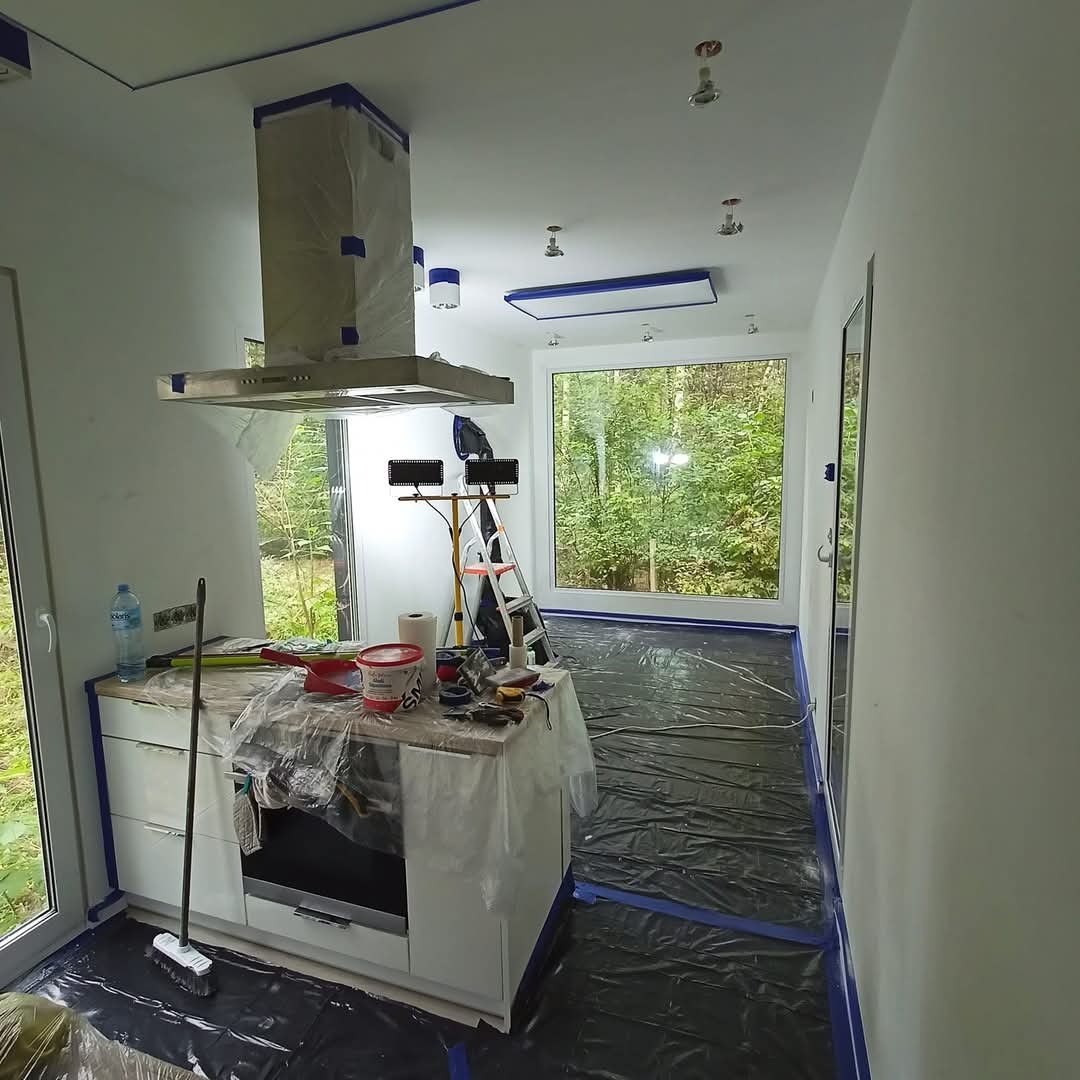 Przygotowanie kuchni do malowania: zabezpieczone folią meble i podłoga, taśma malarska na oknach i okapie. Widoczne narzędzia i drabina. Duże okno z widokiem na zieleń.