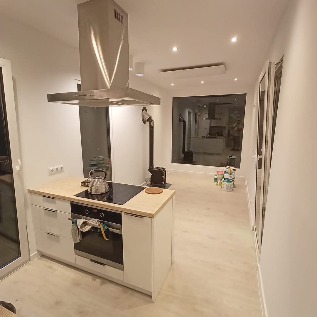 Jasne wnętrze po malowaniu: białe ściany, wyspa kuchenna z płytą, okno z widokiem na dalszą część domu. Na podłodze widoczne puszki z farbą.